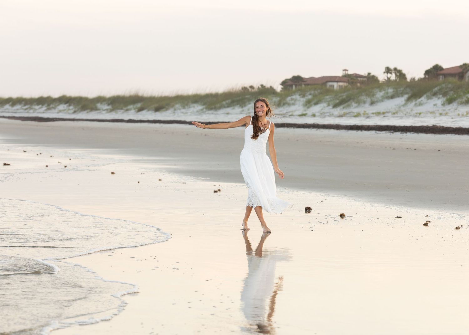 High school senior girl walking along the beach with her arm out for balance as she laughs at the camera.