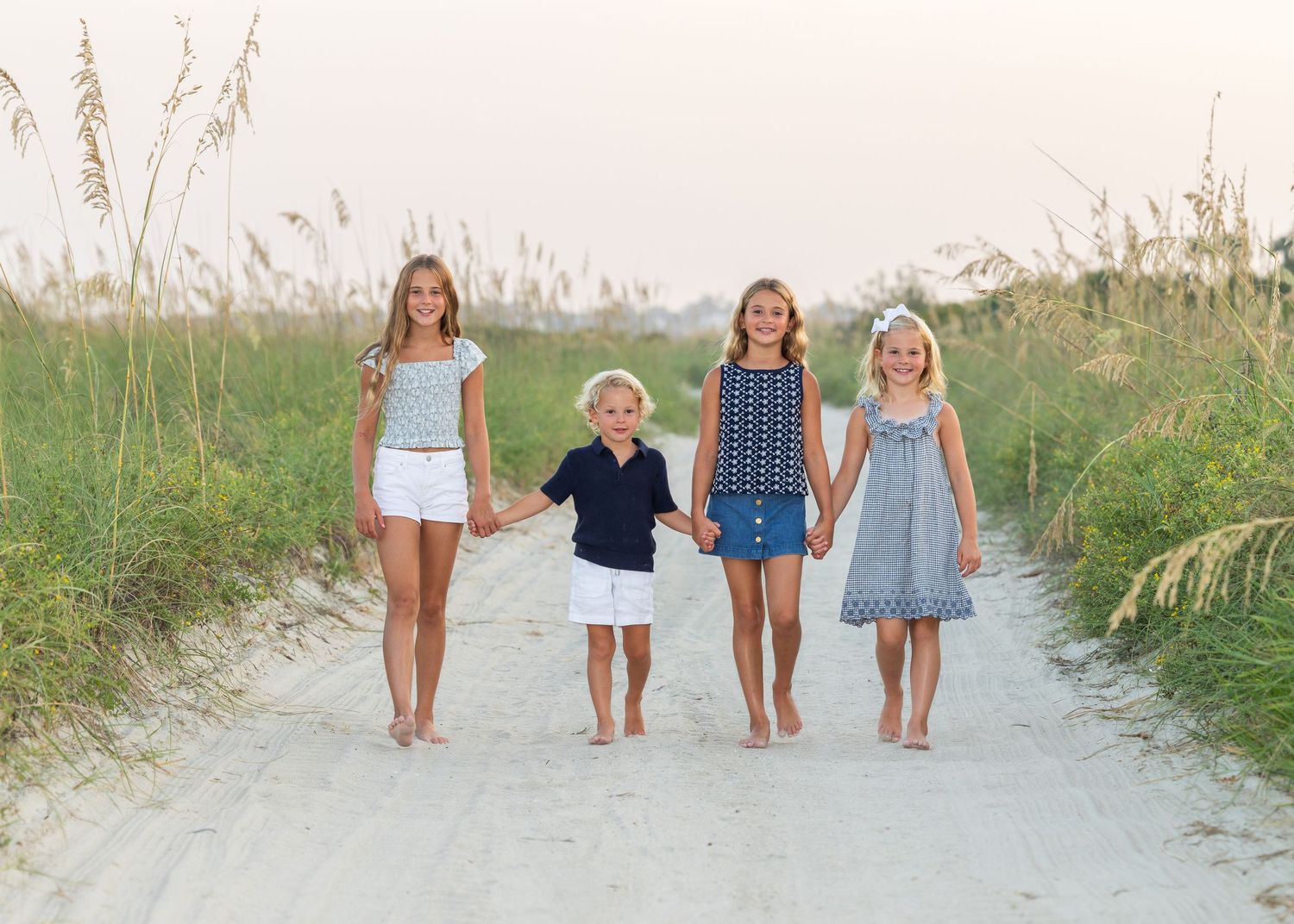 4 siblings walking towards the camera, smiling on a sandy road surrounded by dunes and oats.
