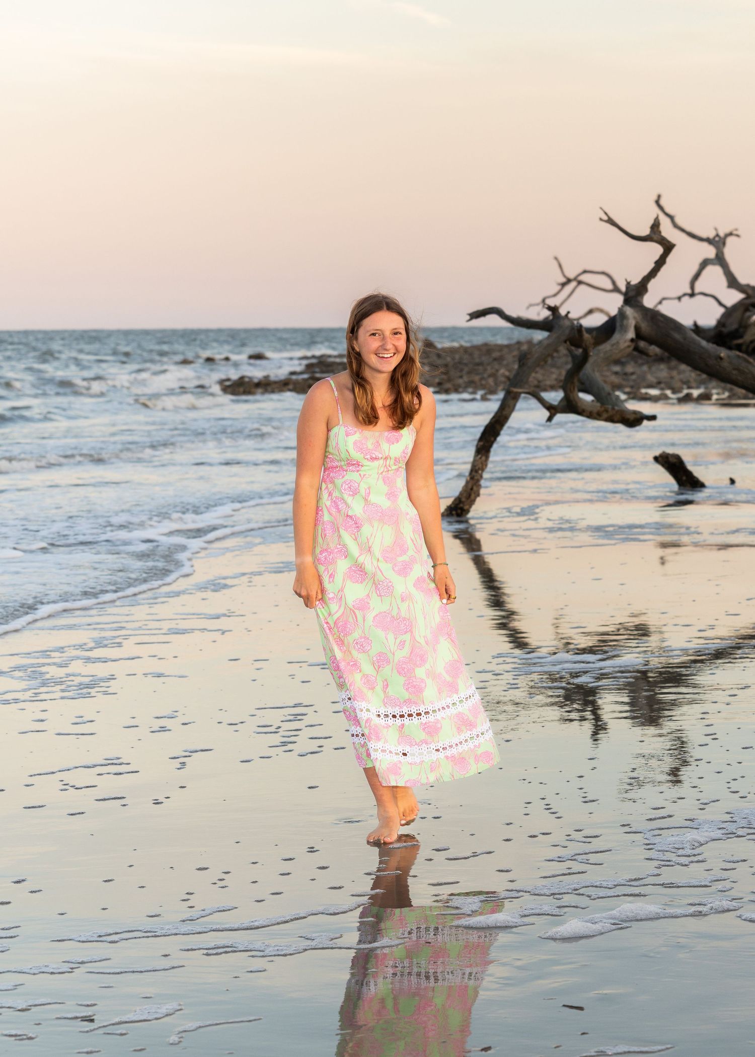 A high school senior girl in a long, flowy dress walking in the water at Driftwood Beach while smiling at the camera.