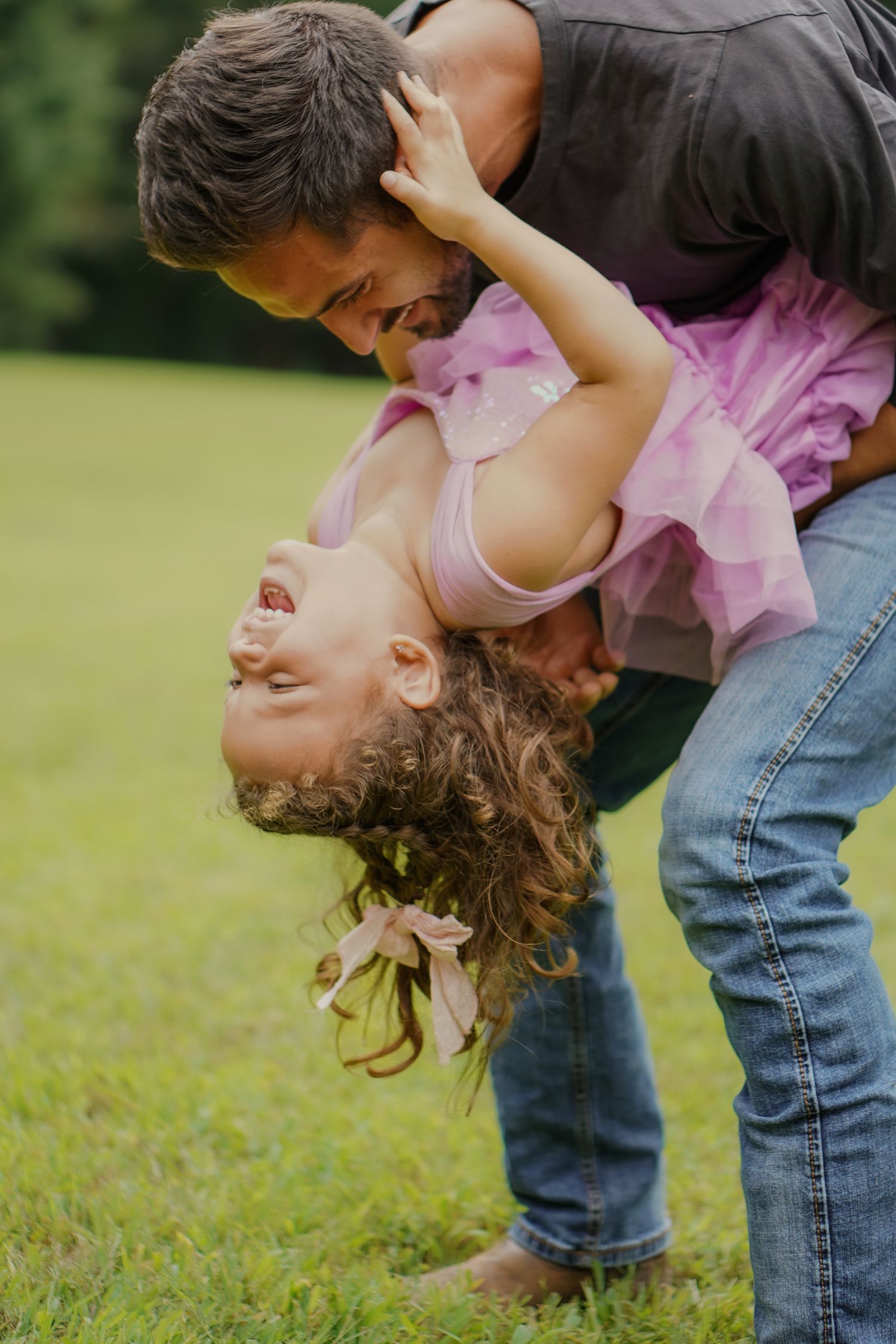Father playfully holds laughing daughter upside down in a green park, girl wearing a pink tutu dress.