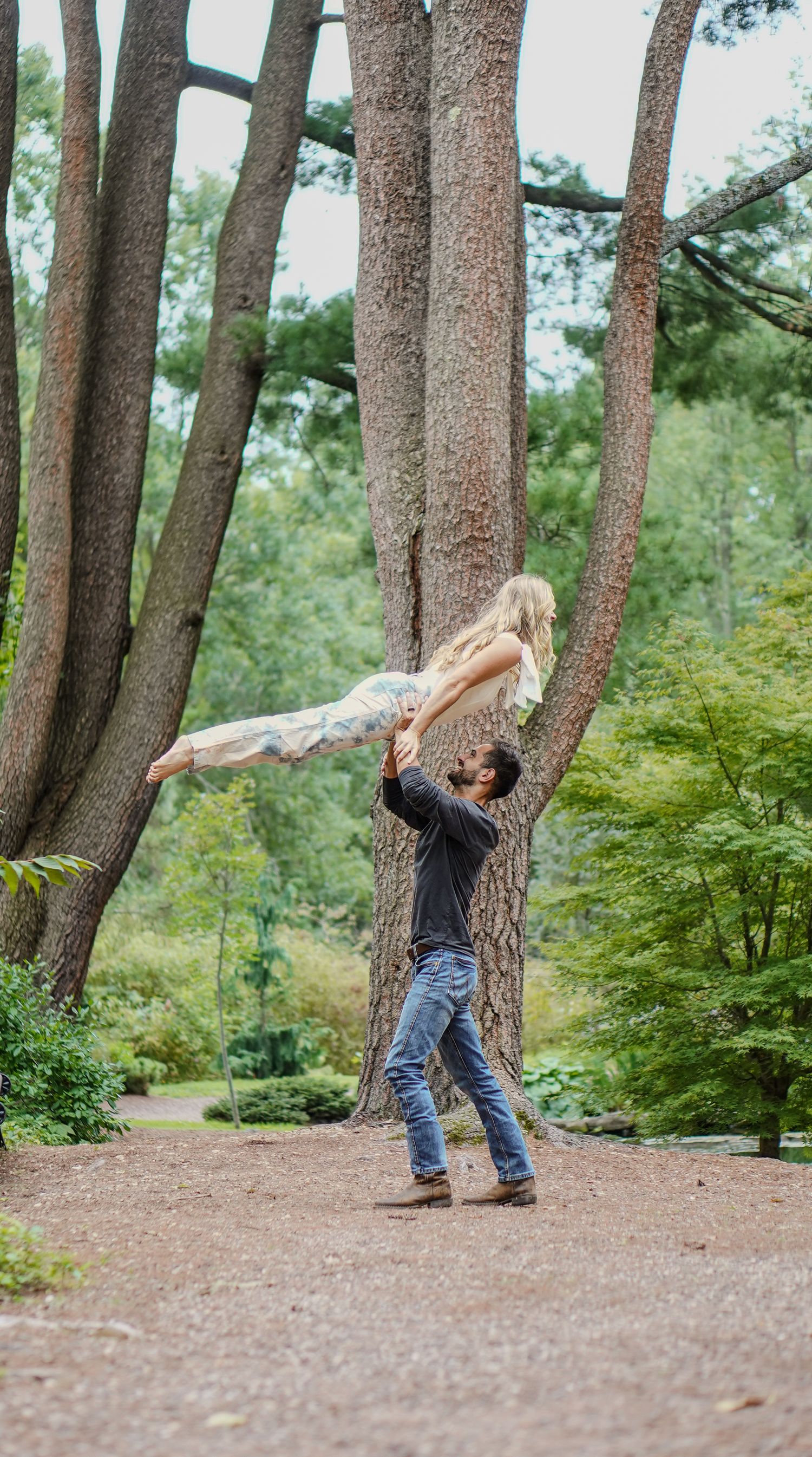 Man dramatically lifts woman high above his head on a tree-lined gravel path in a lush park setting.