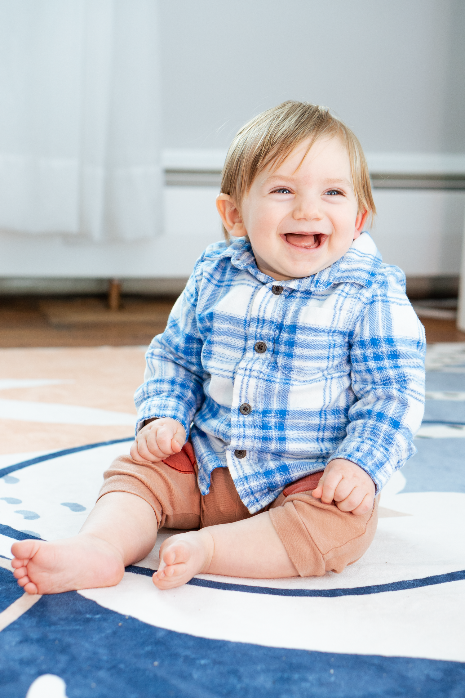 Happy baby boy in blue plaid shirt laughing while sitting on a decorative floor rug indoors.