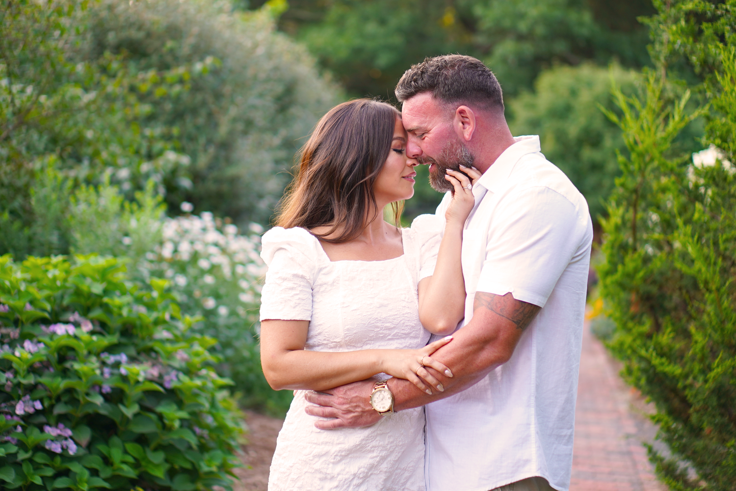 Couple almost kissing while embracing on a garden pathway surrounded by lush green shrubs and flowers.