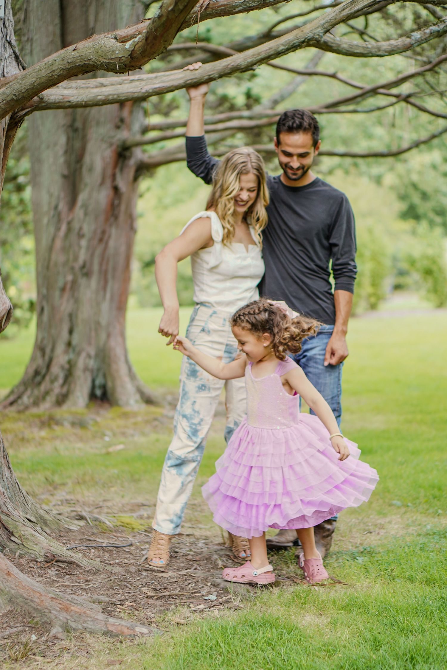 Family of three plays together under a large tree in a green park, young girl in pink tutu dress.