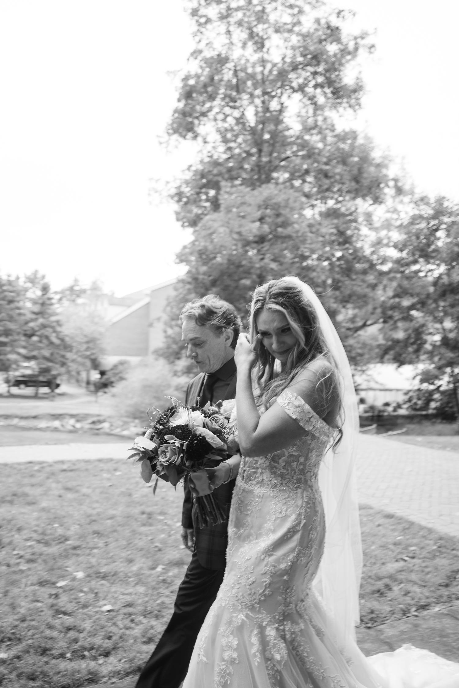 Black and white photo of a bride in lace gown and veil walking with a man outdoors on a wedding day.