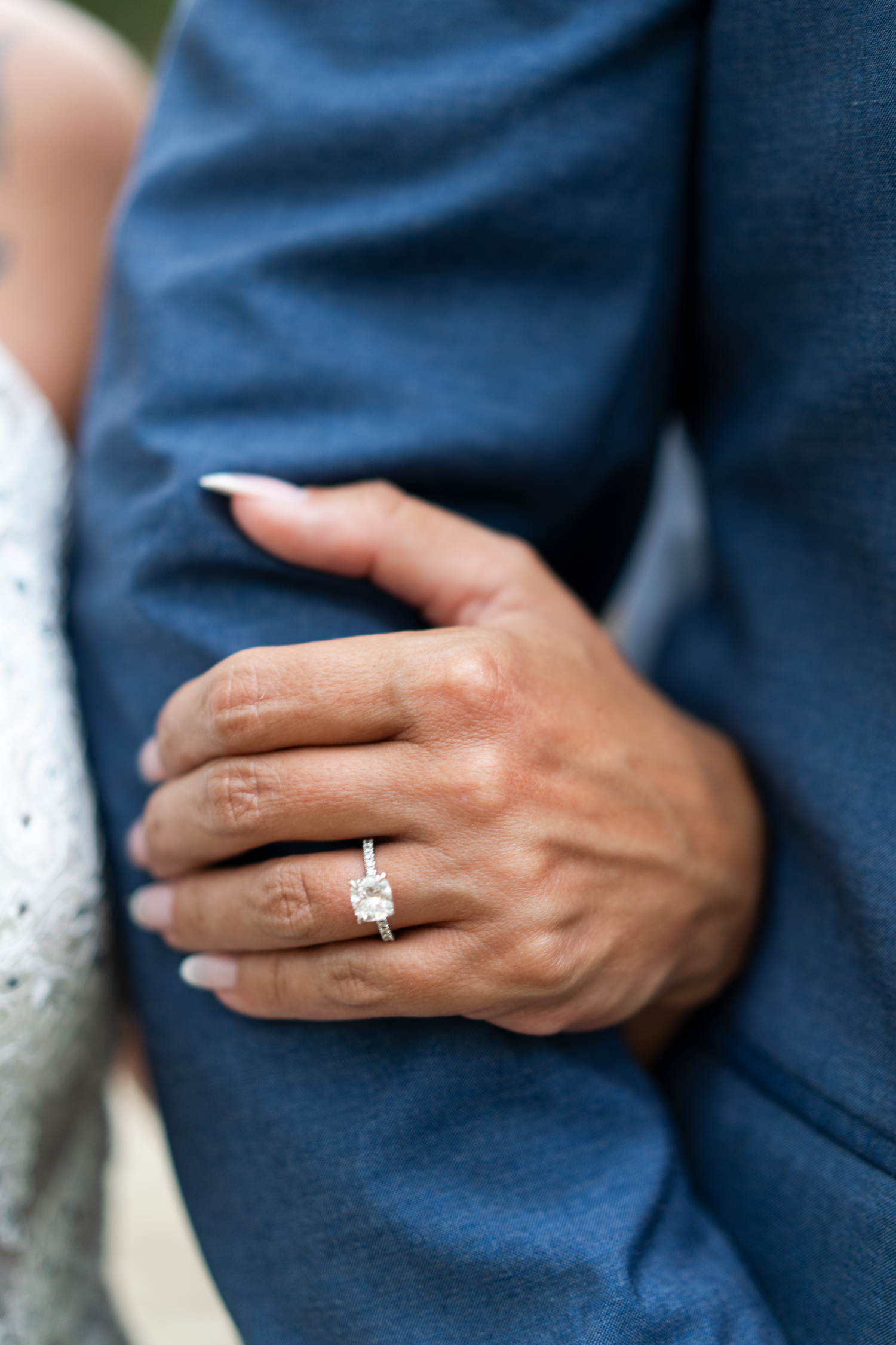 Close-up of bride's hand with oval diamond solitaire engagement ring resting on groom's navy sleeve.