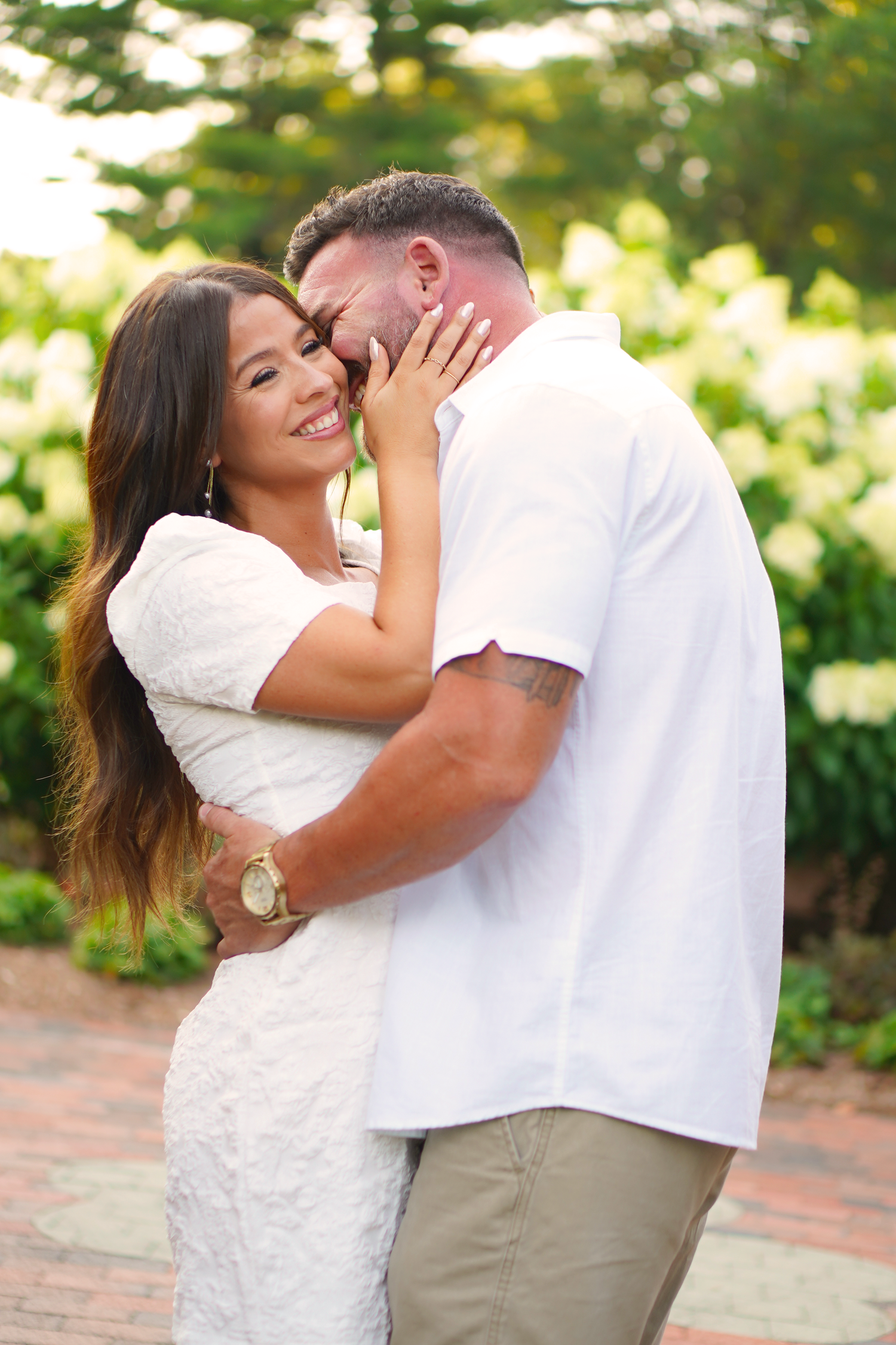 Smiling couple embracing outdoors, woman in white lace dress holds mans face as they share a romantic moment in a garden.