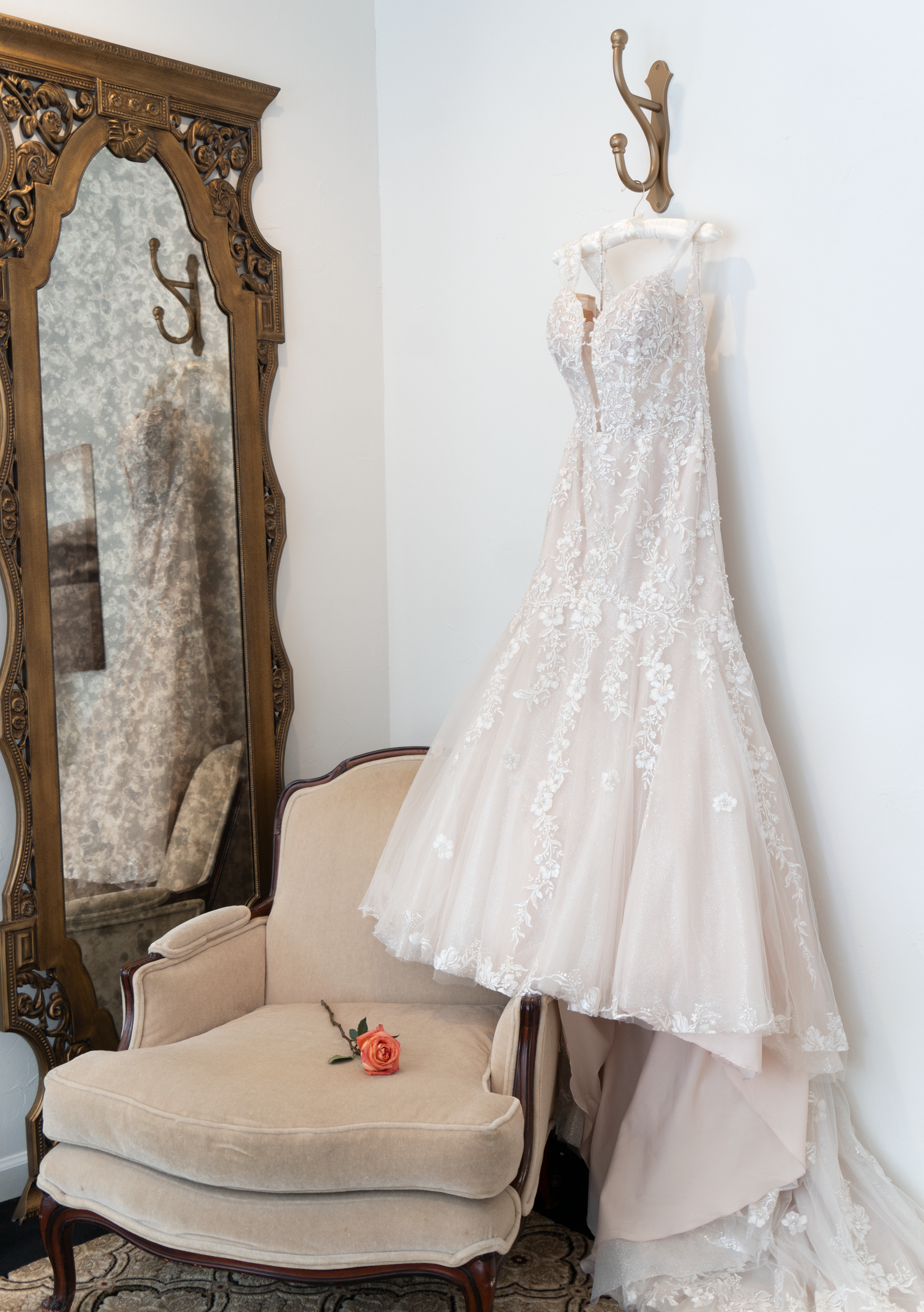Elegant lace wedding gown hanging on wall hooks beside an ornate mirror and vintage chair with a pink rose.