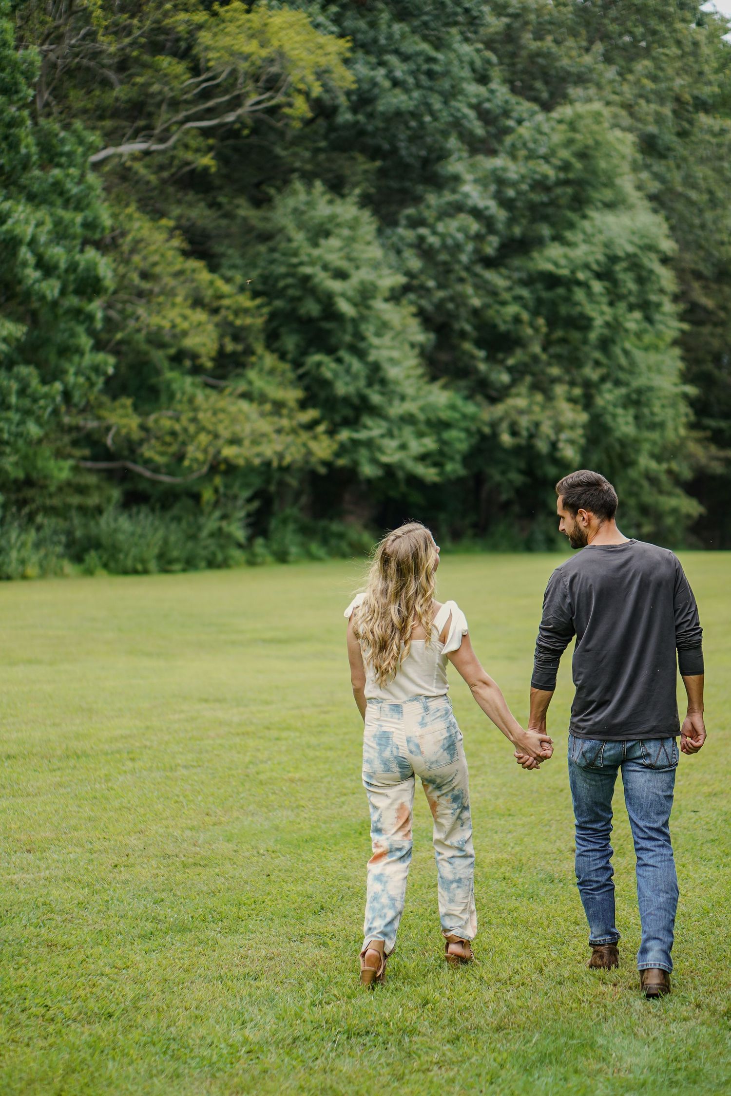 Couple holding hands walking away through a lush green field toward a tree line on a cloudy day.
