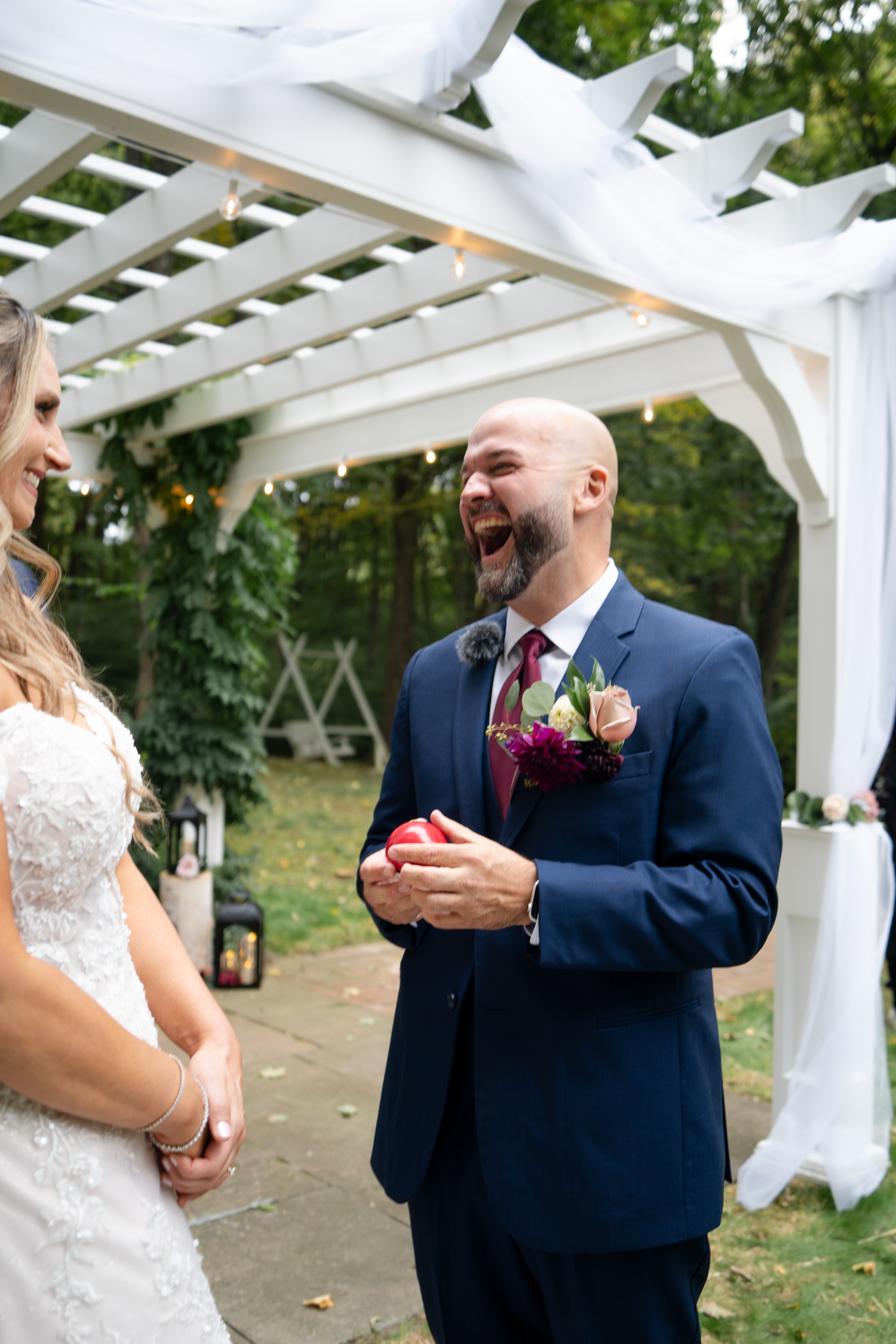 Laughing bald groom in navy suit holding ring box at outdoor wedding ceremony under white pergola.