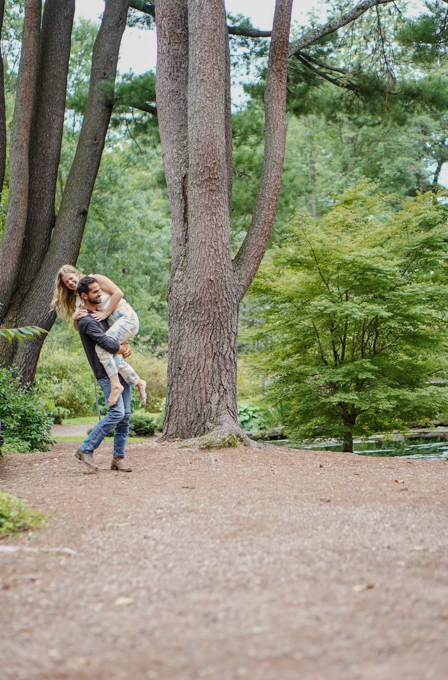 Couple sharing a joyful embrace on a wooded garden path surrounded by tall trees and green foliage.