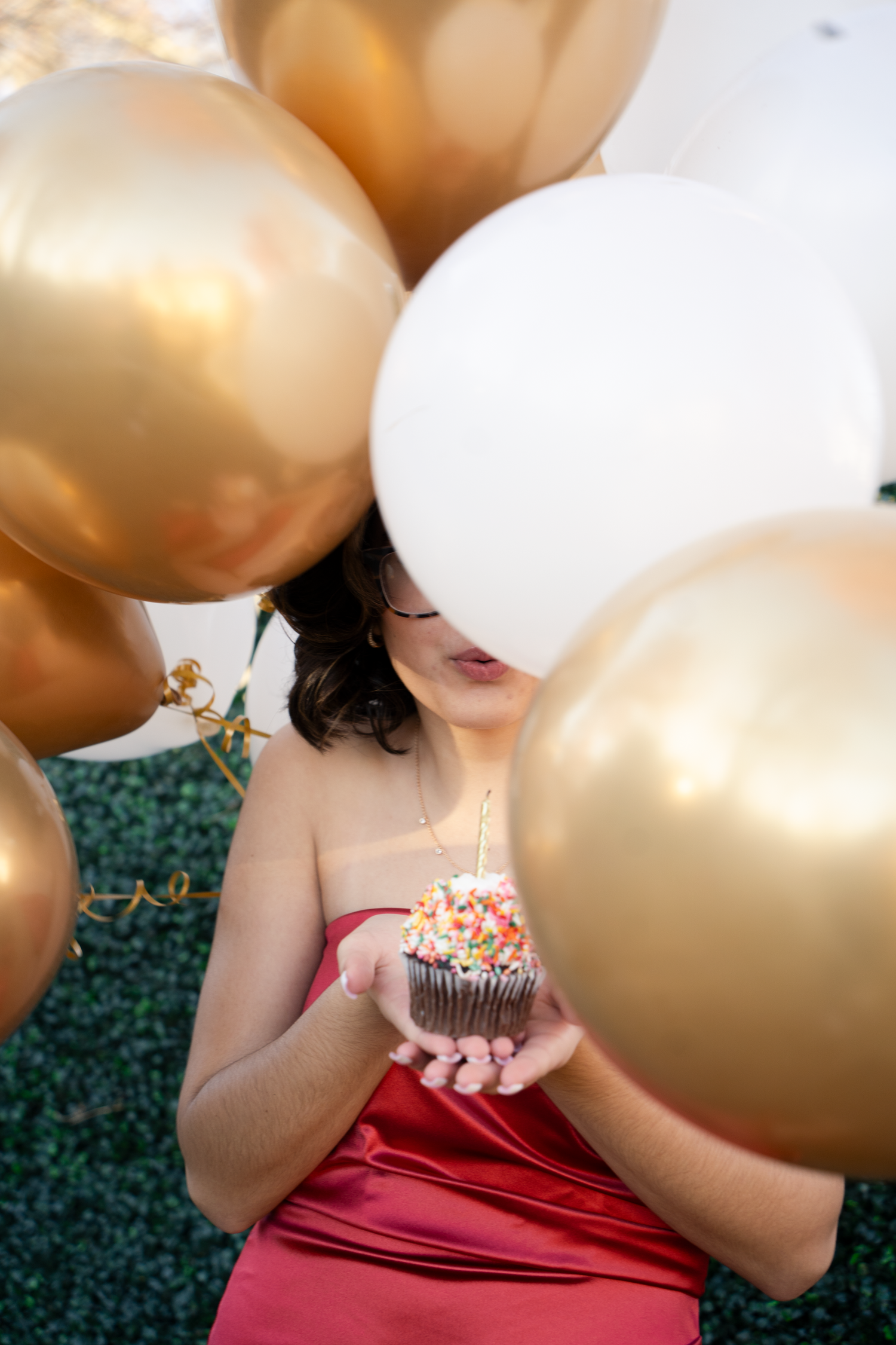 Woman in red dress blowing out candle on sprinkle cupcake surrounded by gold and white balloons.