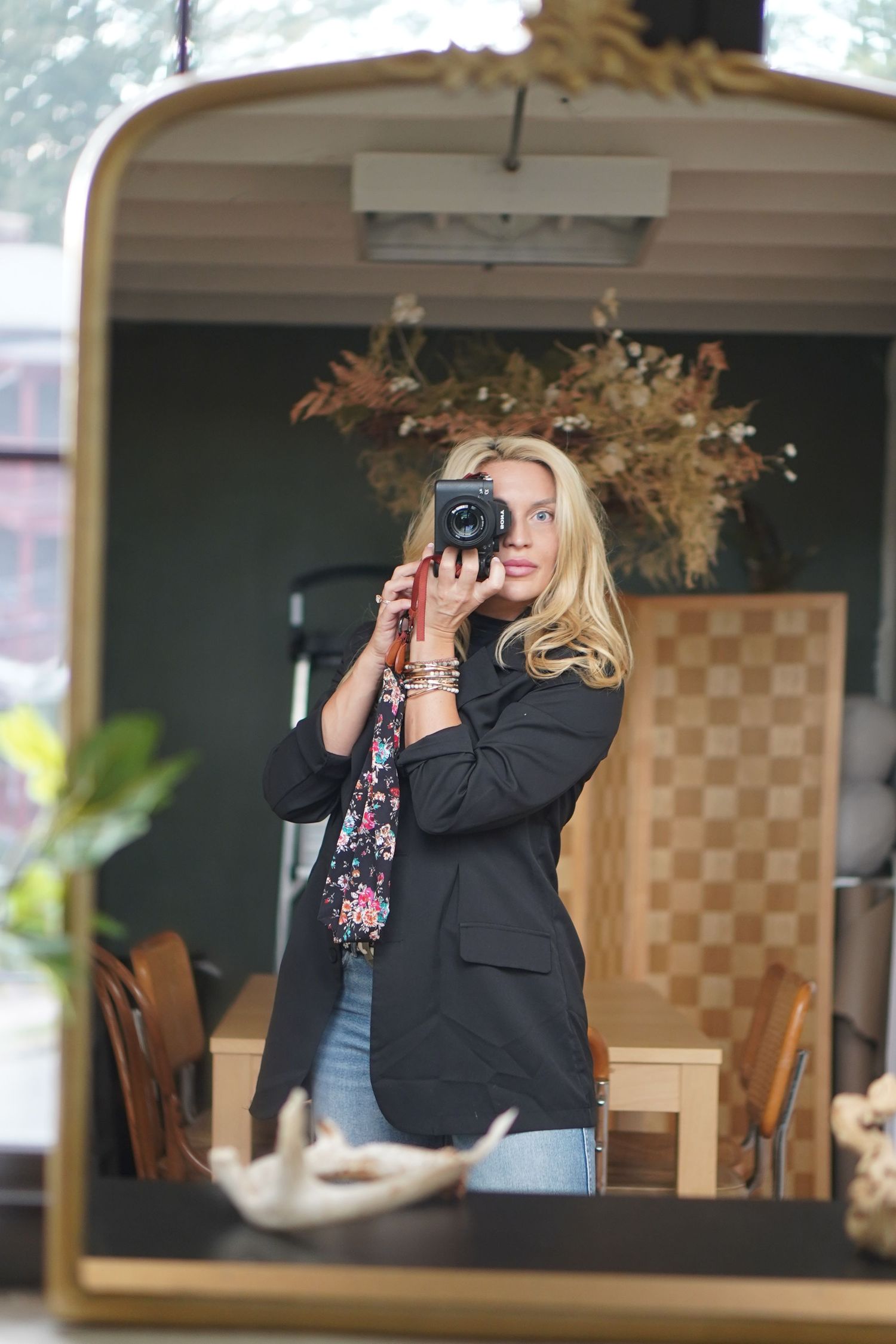 Blonde woman taking a mirror selfie with a black camera, wearing a navy blazer in a cozy cafe setting.
