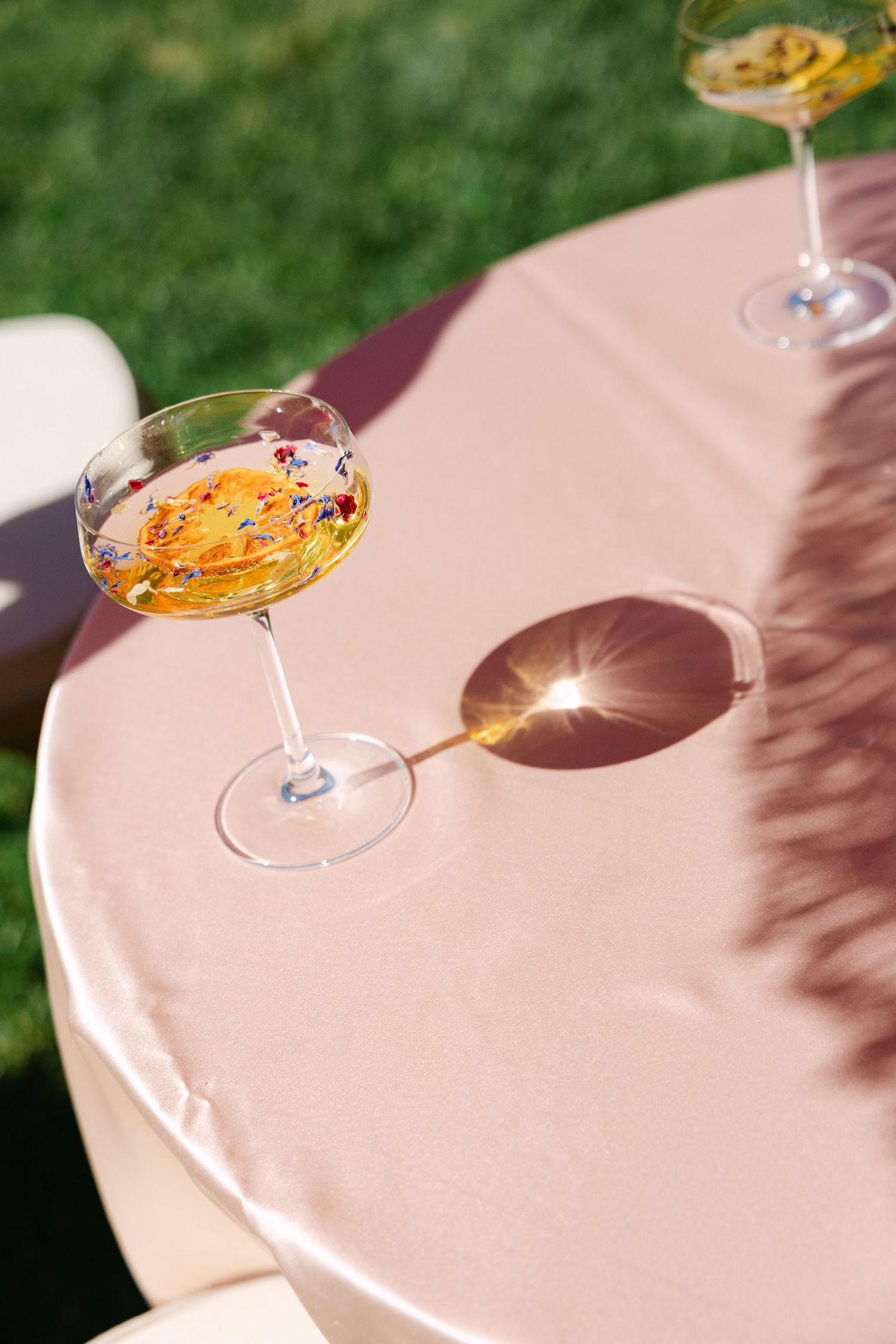 Two elegant coupe glasses with floral garnishes sit on a blush pink tablecloth in bright sunlight outdoors.