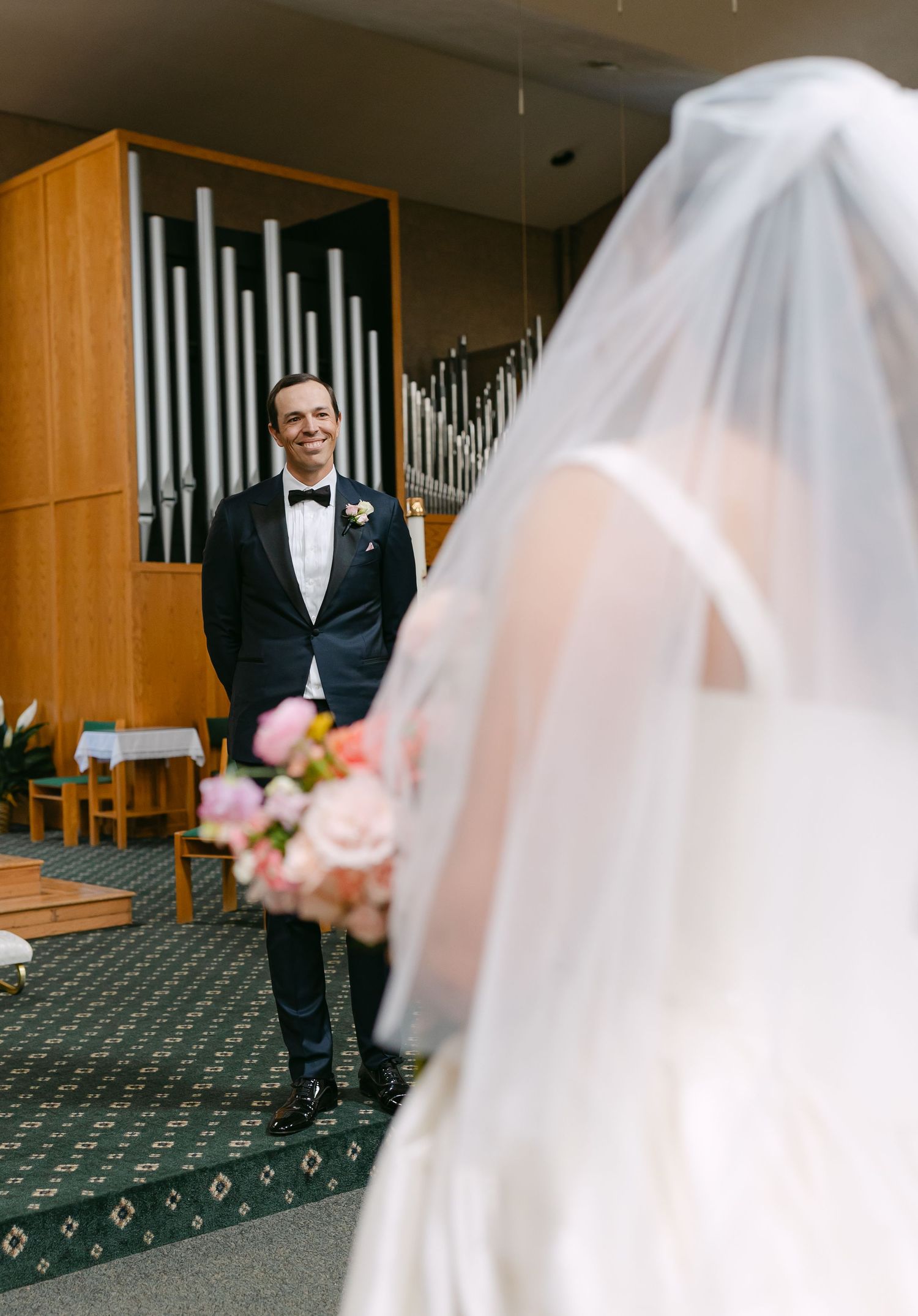 Groom in tuxedo smiling at bride in white wedding gown and veil inside a church with pipe organ backdrop.