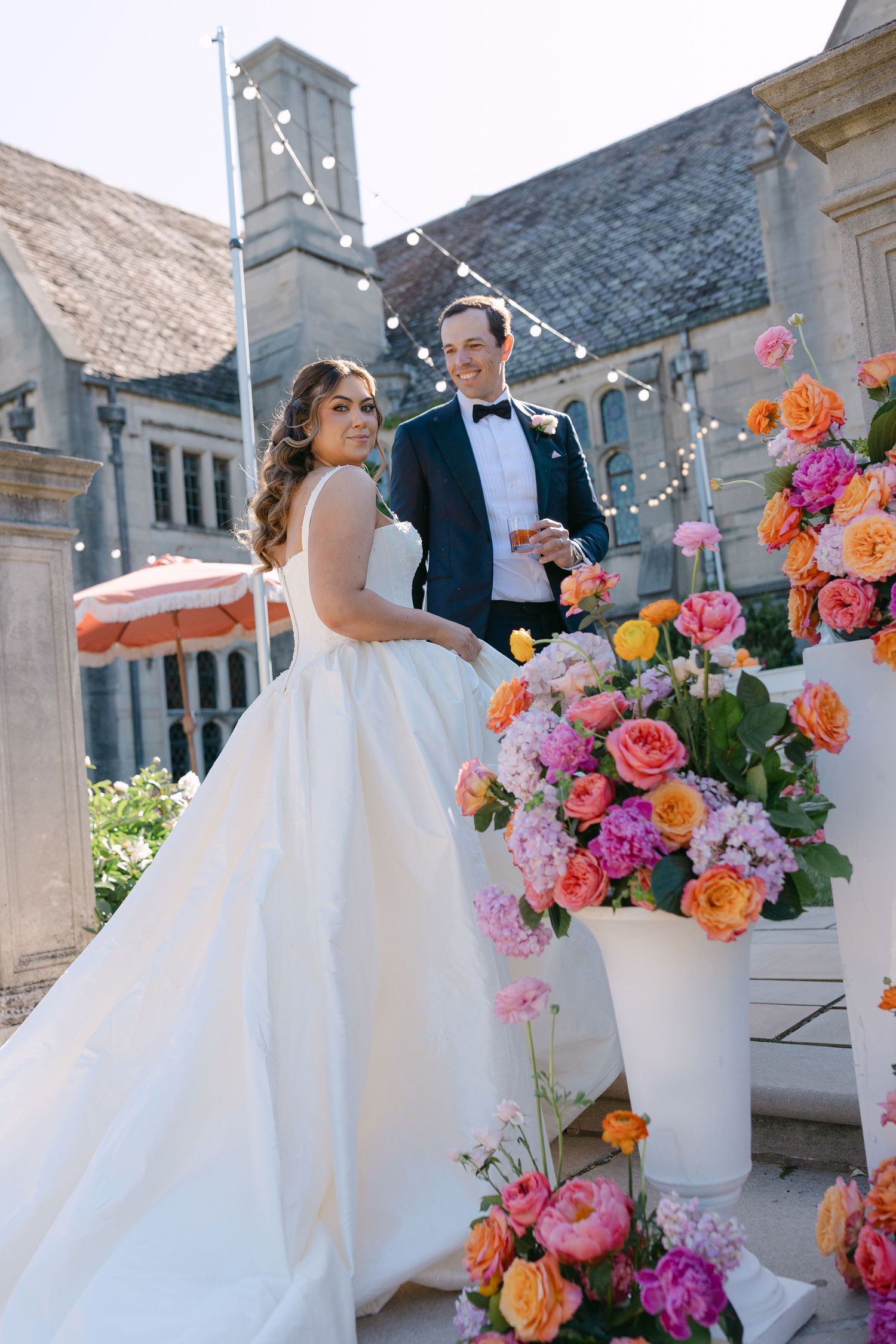 Bride and groom stand beside tall arrangements of colorful orange and pink flowers at venue.