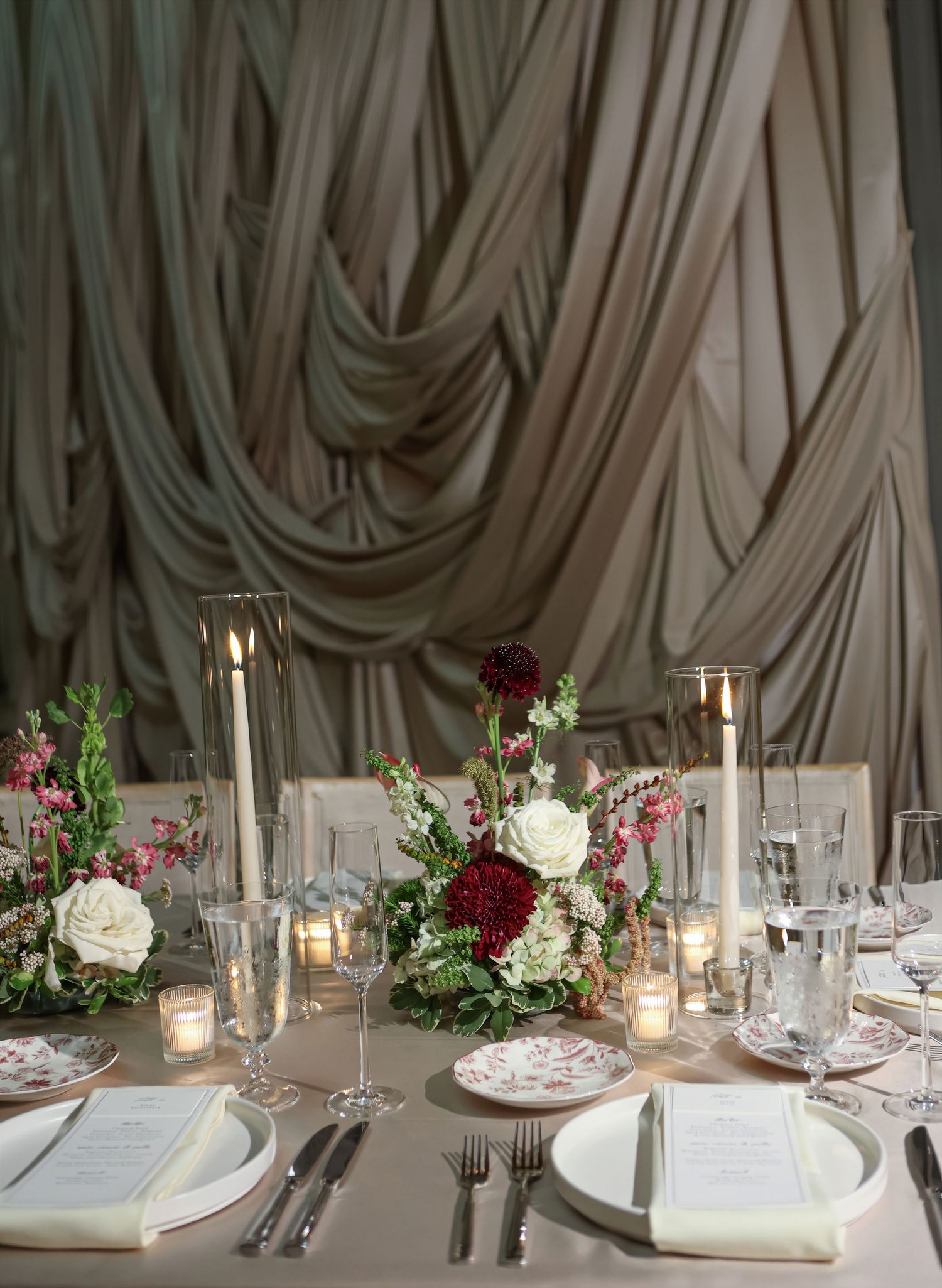 Romantic wedding tablescape with white and burgundy florals, tall candles, and draped fabric backdrop.