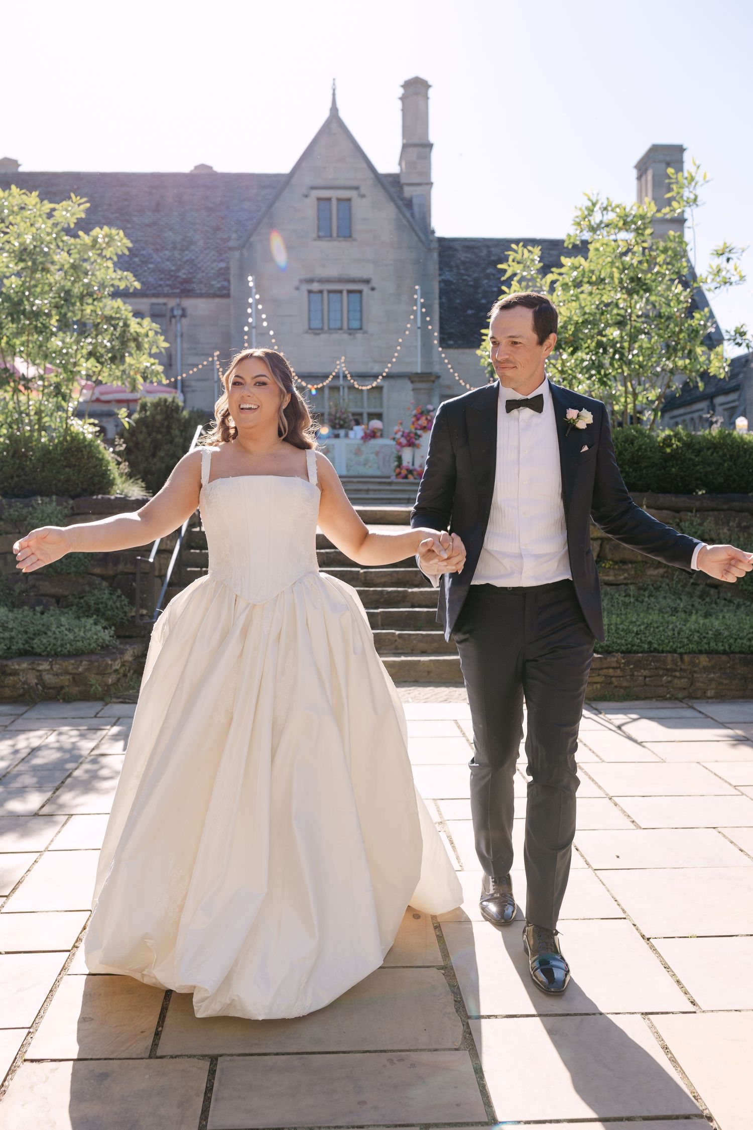 Happy bride and groom holding hands walking on stone path outside a historic manor on their wedding day.