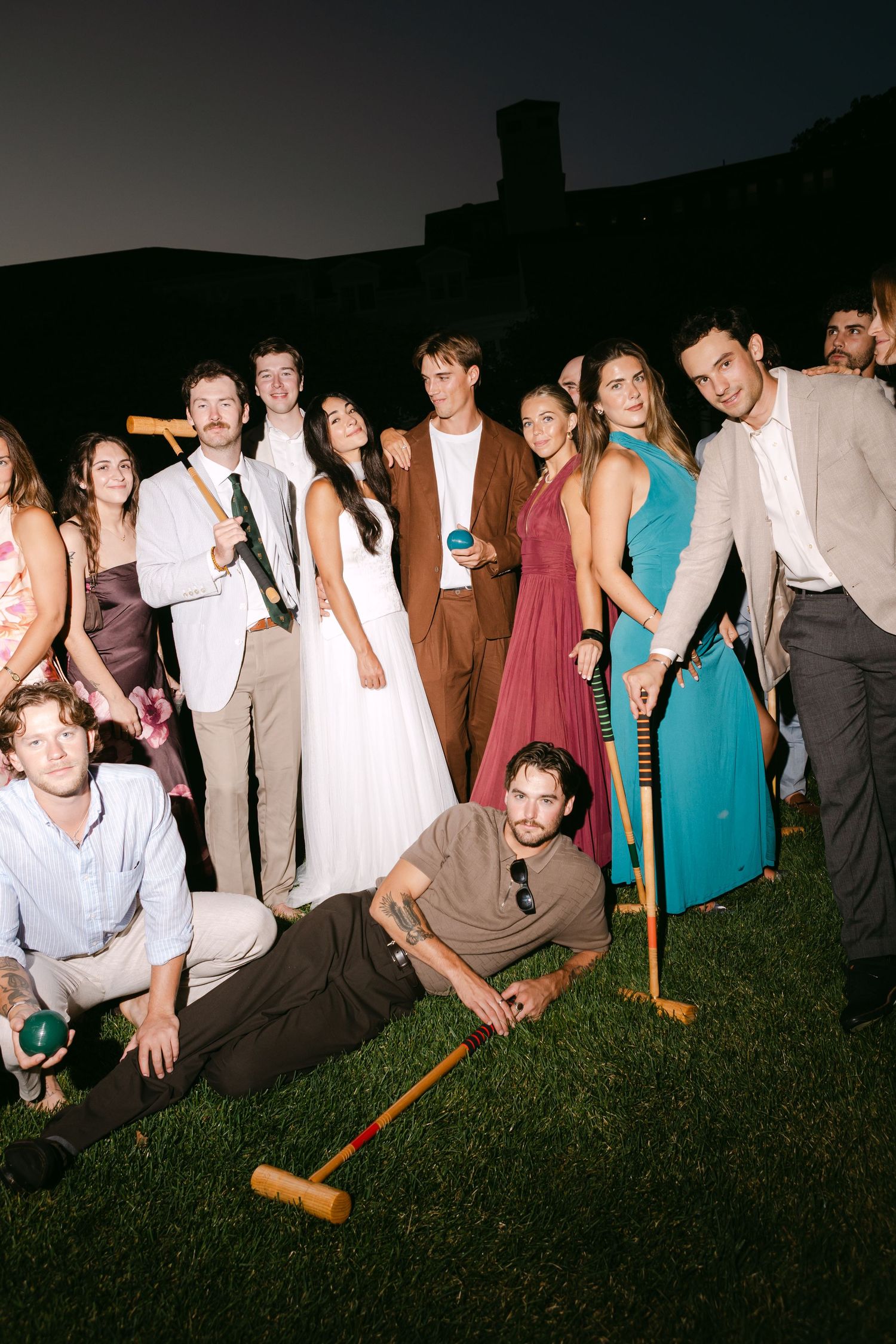 A group of elegantly dressed wedding guests pose outdoors at night, holding croquet mallets on a lawn.
