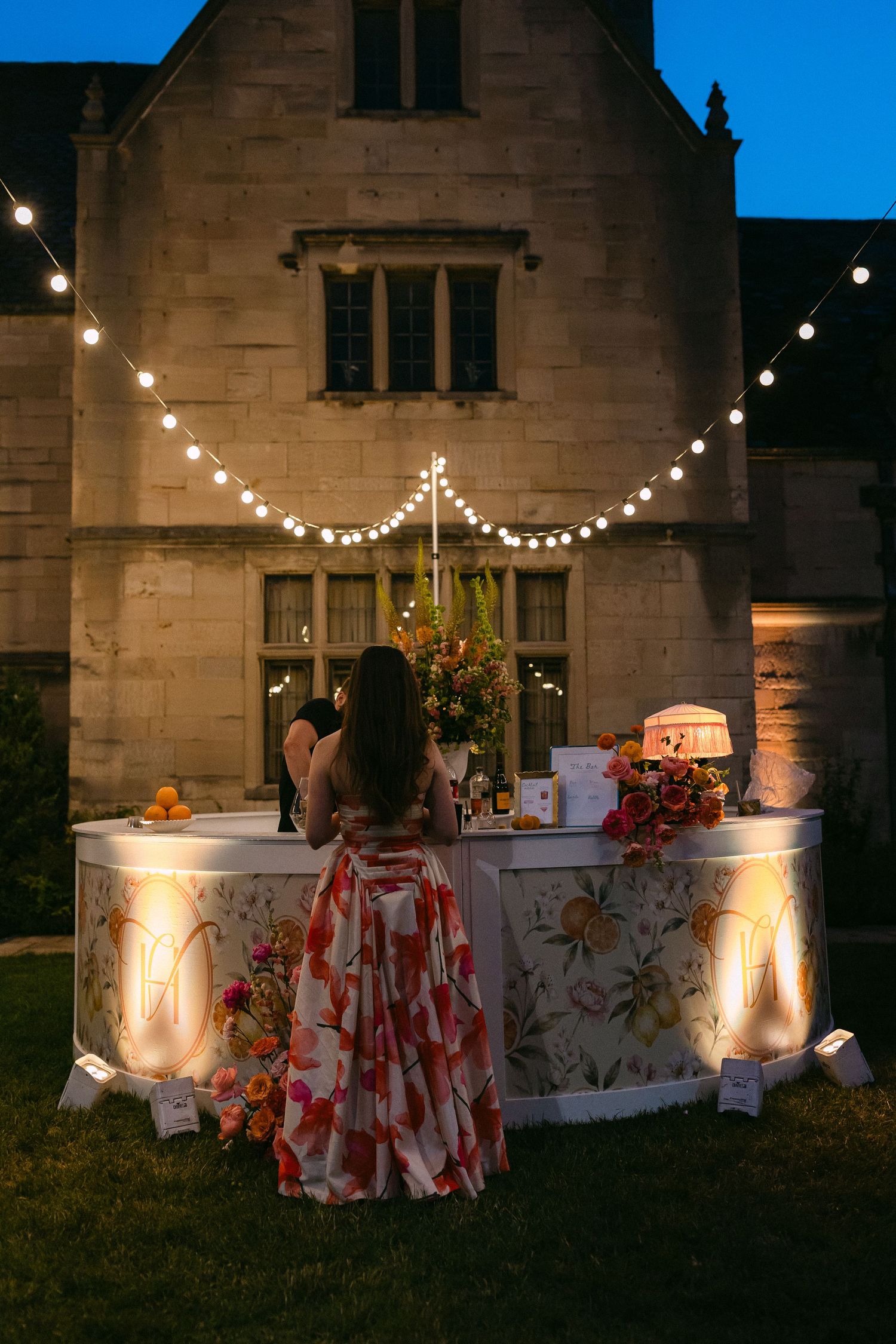 Woman in floral gown standing at illuminated outdoor wedding bar beneath string lights at night.