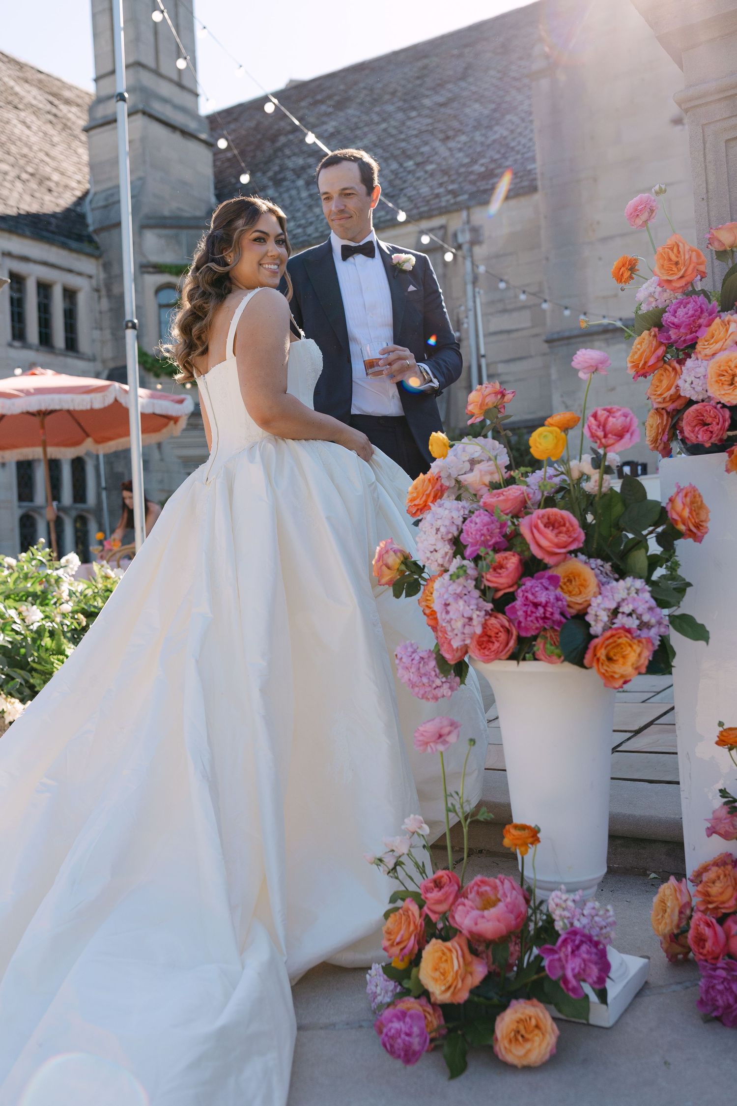 Bride and groom posing outdoors near vibrant pink and orange floral arrangements at reception.