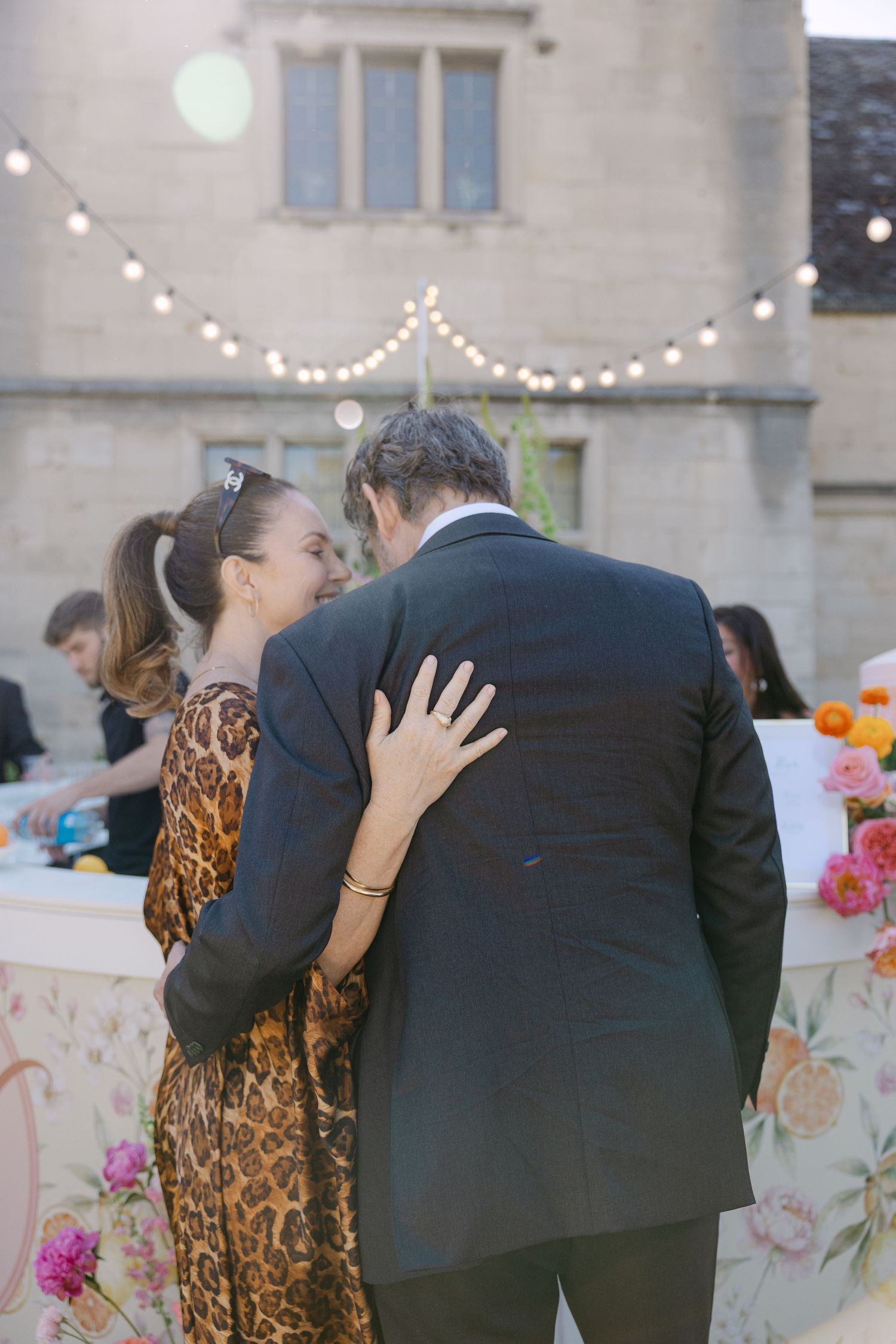 Couple embracing at outdoor wedding reception with string lights and floral bar backdrop at dusk.