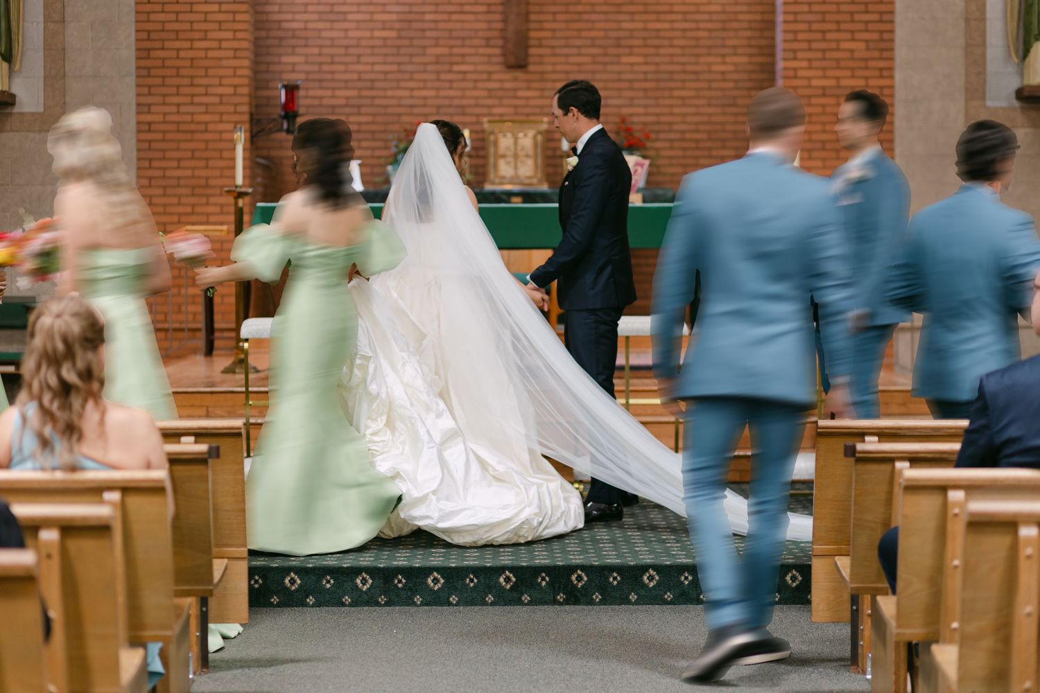 Bride and groom kneeling at church altar during wedding ceremony with bridal party in sage green and blue attire.