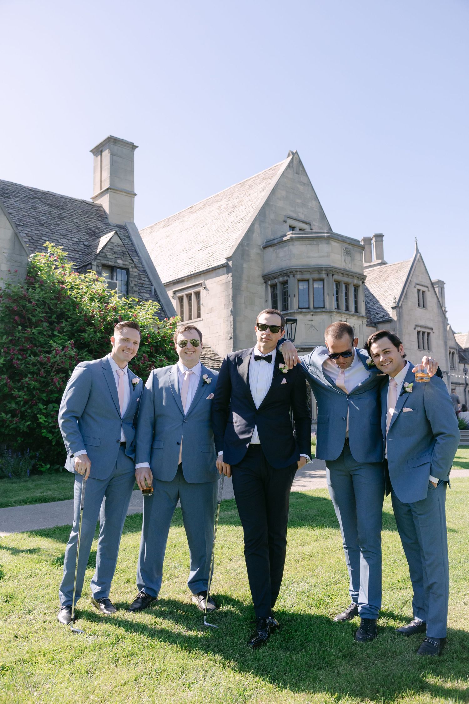 Groom in black tuxedo poses with four groomsmen in light blue suits outside elegant stone manor.