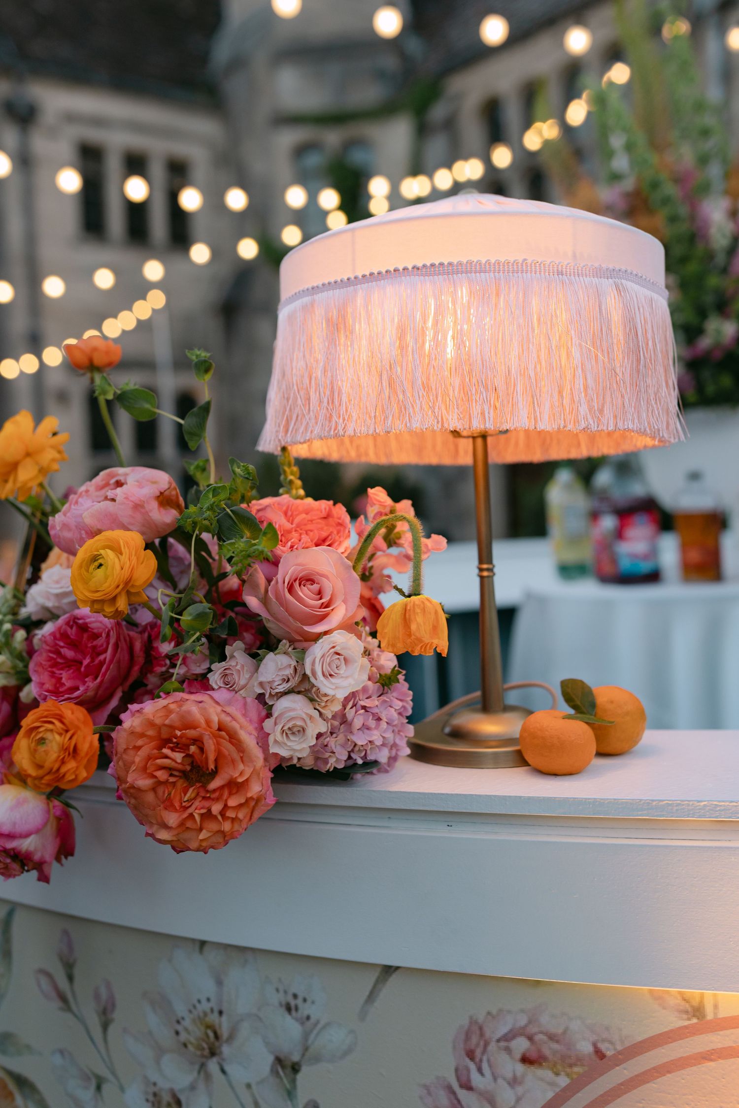 Elegant wedding bar detail with fringe lamp, colorful ranunculus and rose floral arrangement, and string lights at dusk.