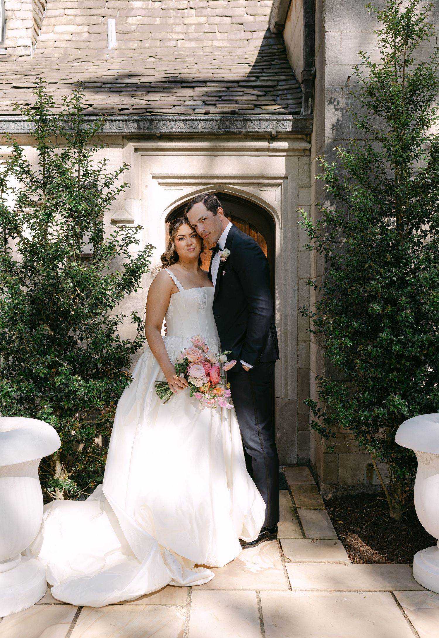 Bride in white ball gown holds pink bouquet beside groom in tuxedo outside stone church.
