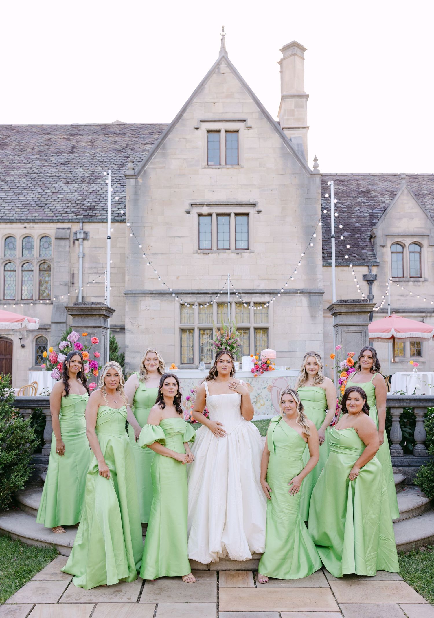 Bride in white gown with bridesmaids in green dresses posing before stone manor wedding venue.
