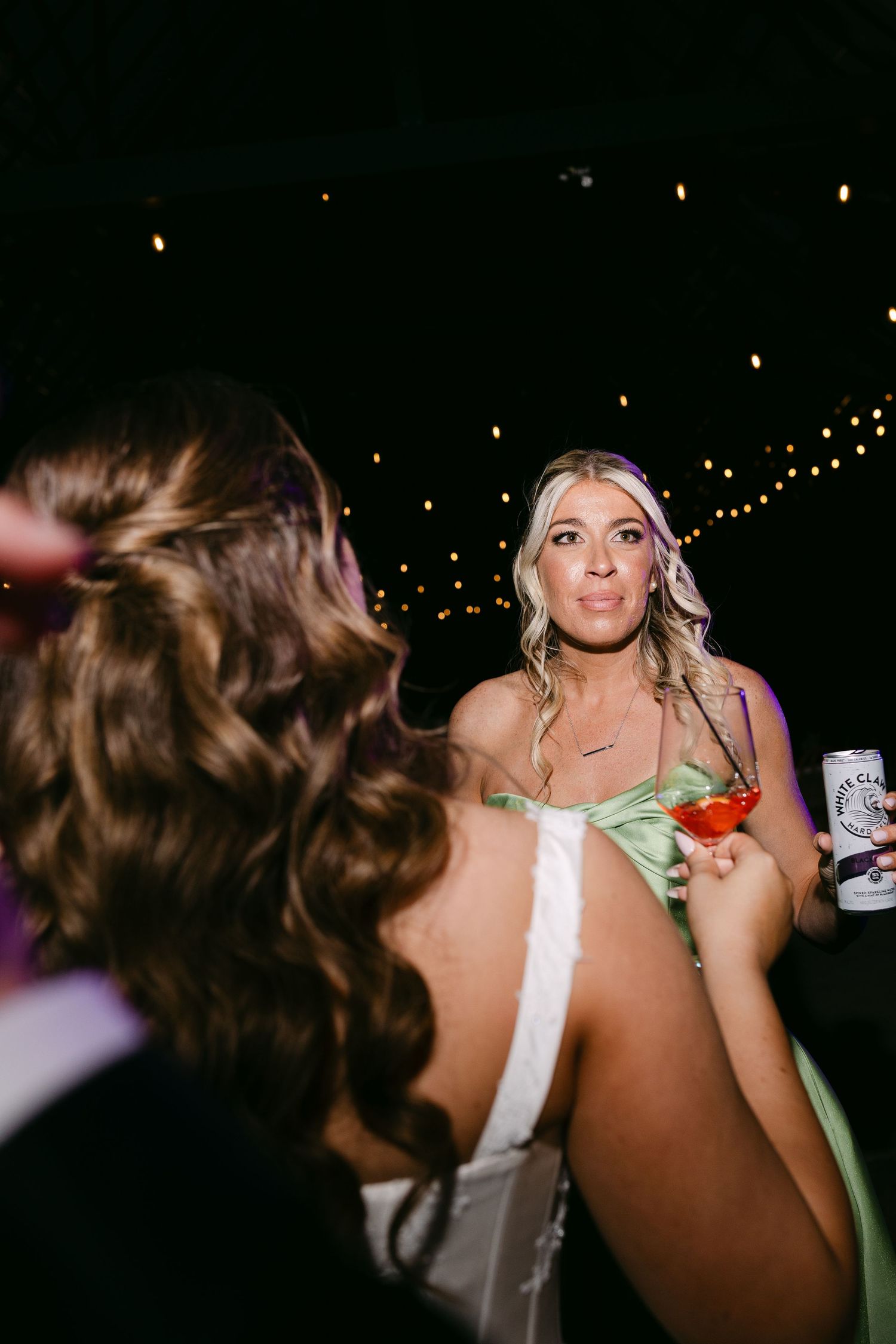 Blonde bride in white gown holds cocktail and beer can while chatting at evening reception.
