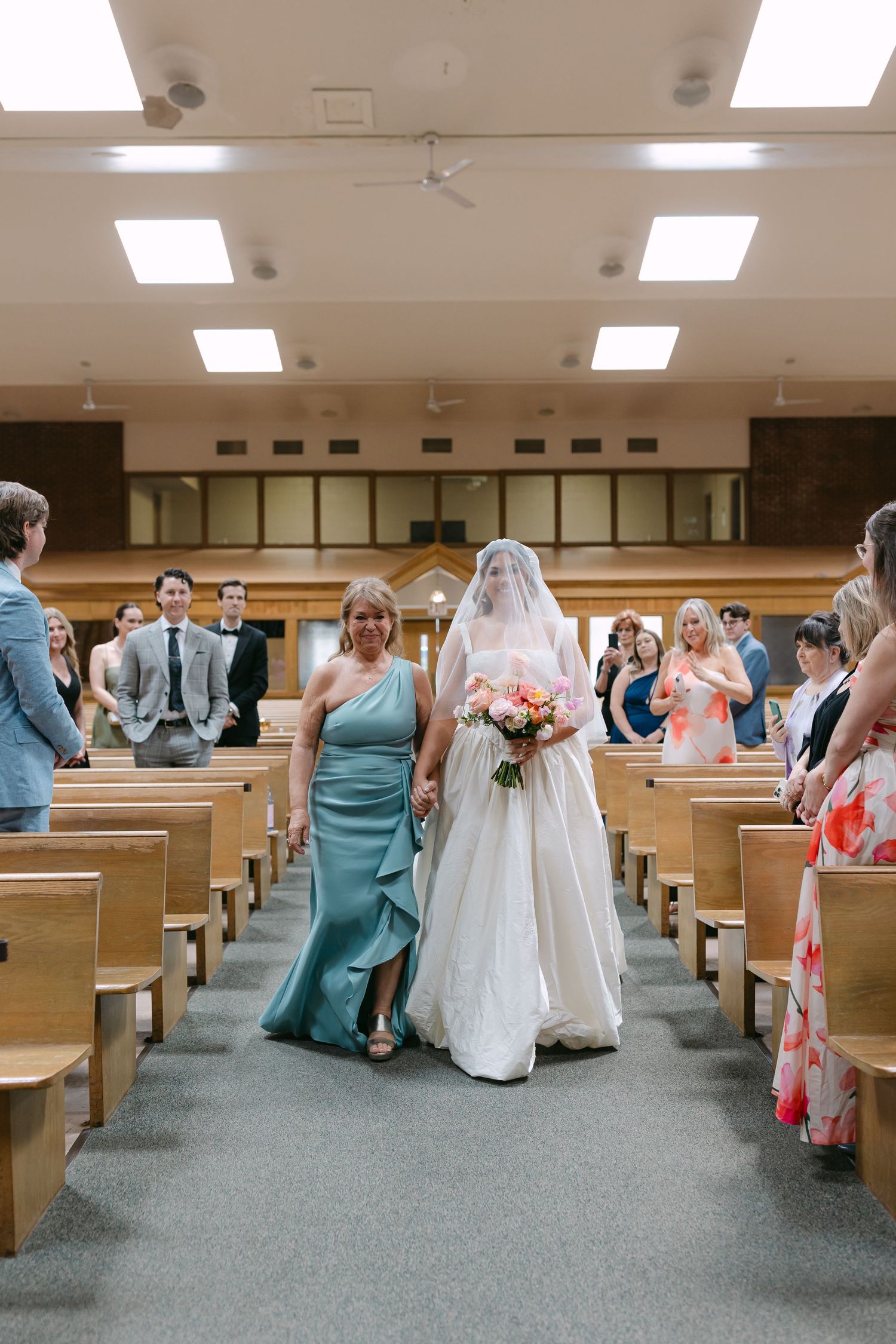 Bride walks down church aisle in white gown and veil, escorted by woman in teal dress, with guests seated in wooden pews.