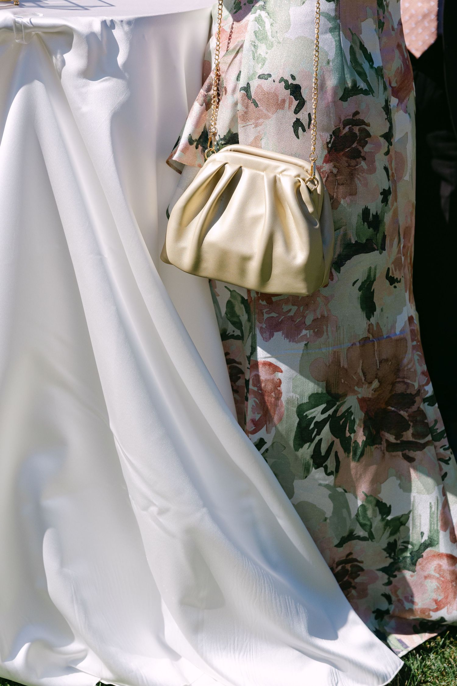 Woman in floral dress holding a gold ruched clutch bag beside a flowing white bridal gown outdoors.