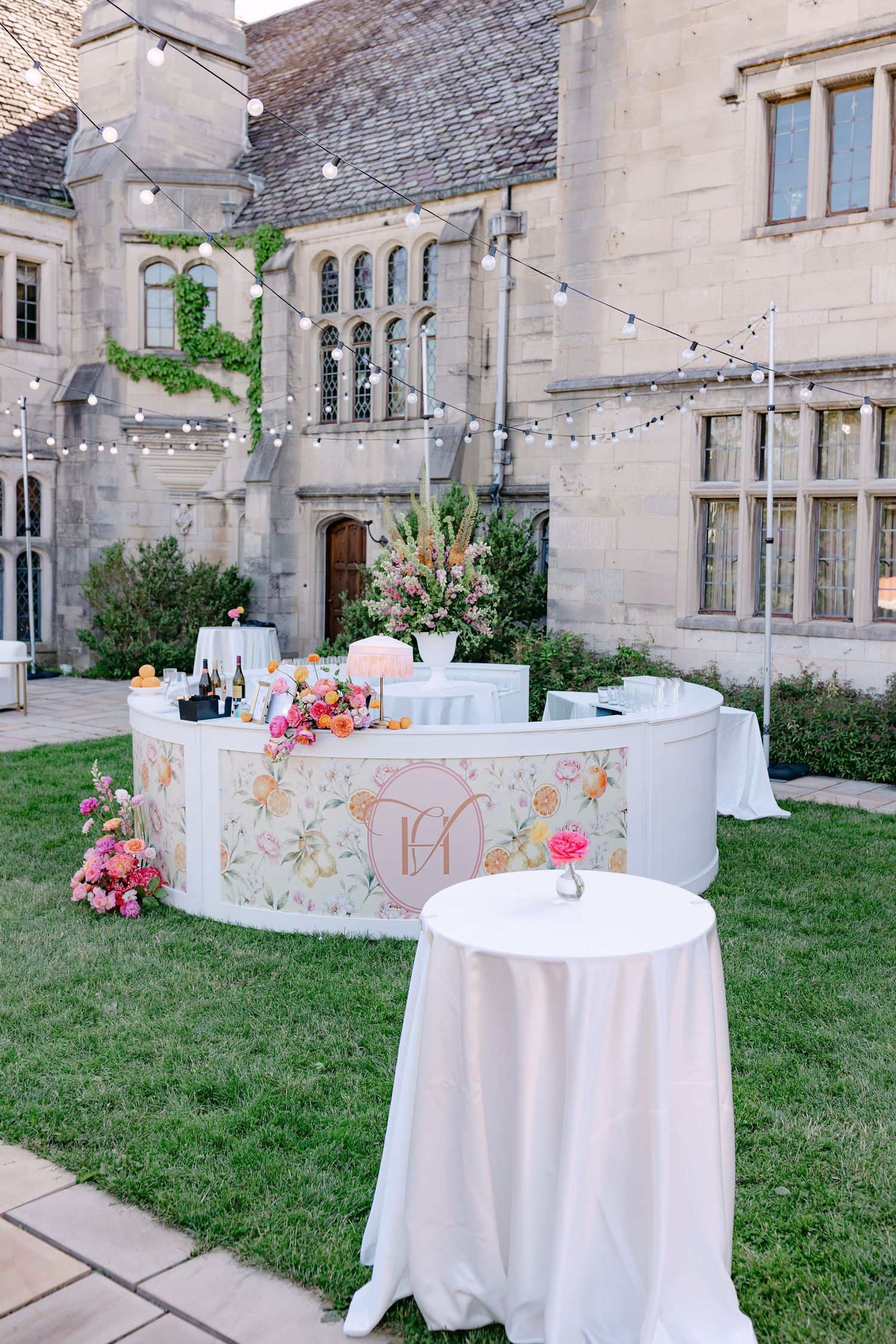 Elegant outdoor wedding bar with floral wrap design, string lights, and colorful flower arrangements at a stone manor.