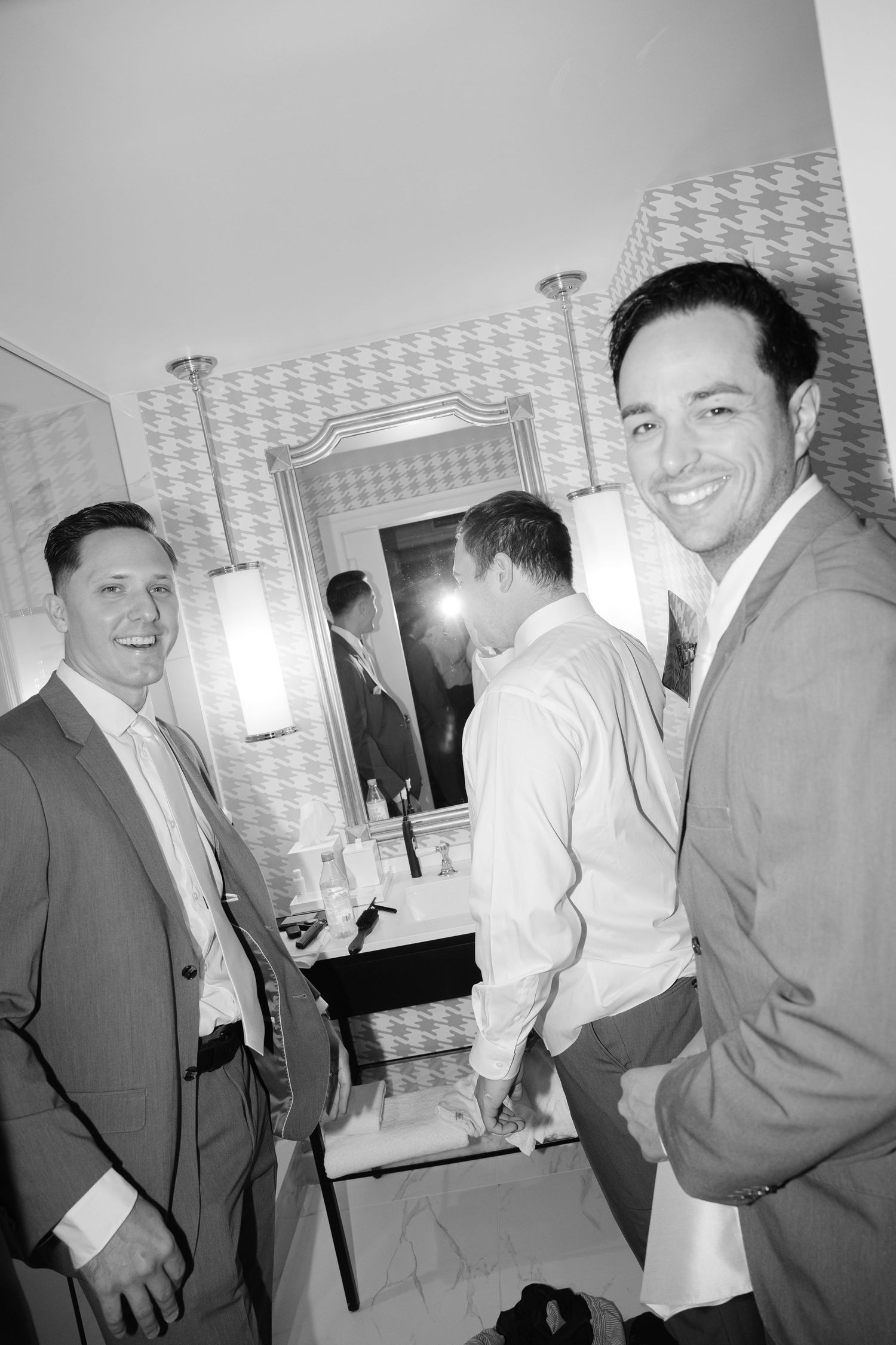 Black and white photo of groomsmen smiling in suits preparing in a hotel bathroom before a wedding.
