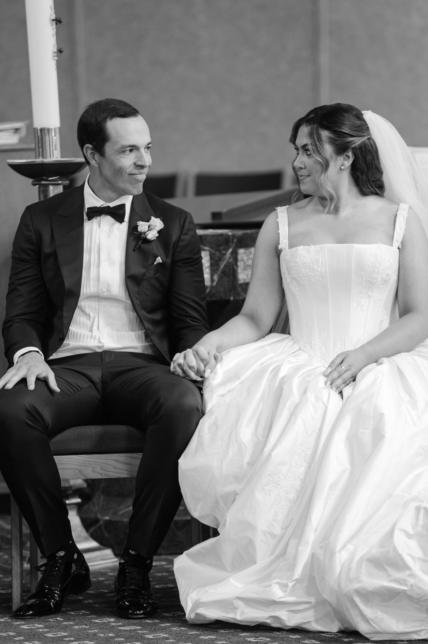 Black and white photo of bride and groom holding hands and smiling at each other during wedding ceremony.