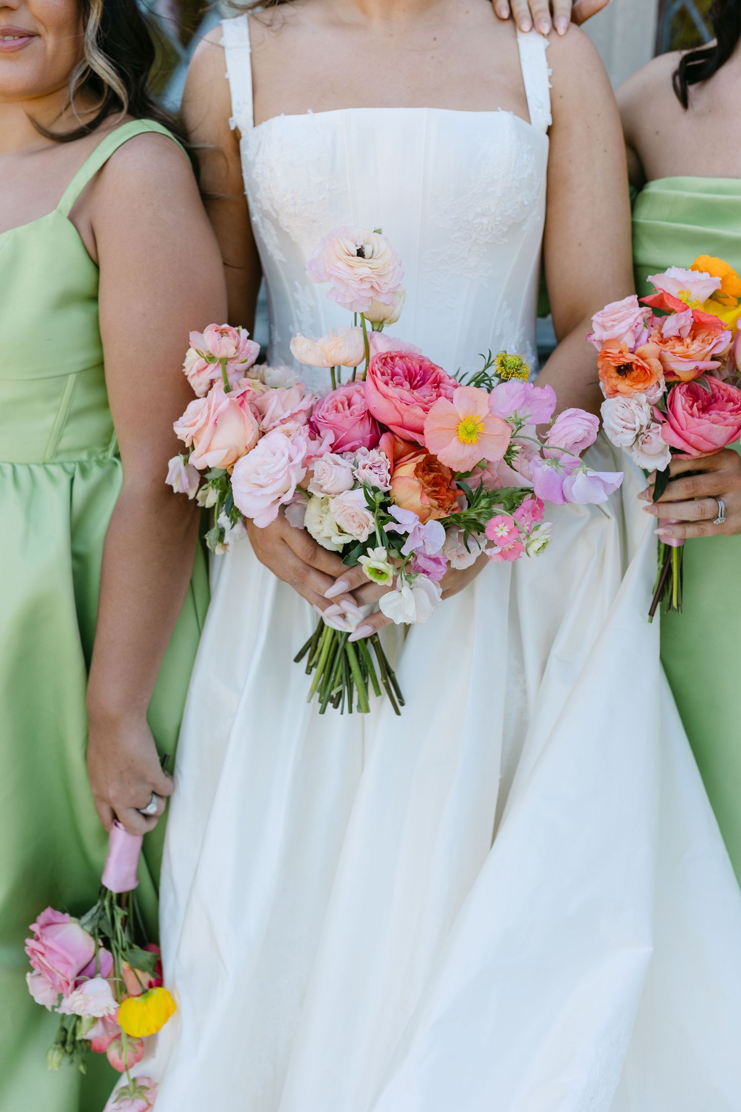 Bride in white gown holding pink and orange bouquet flanked by bridesmaids in green dresses.