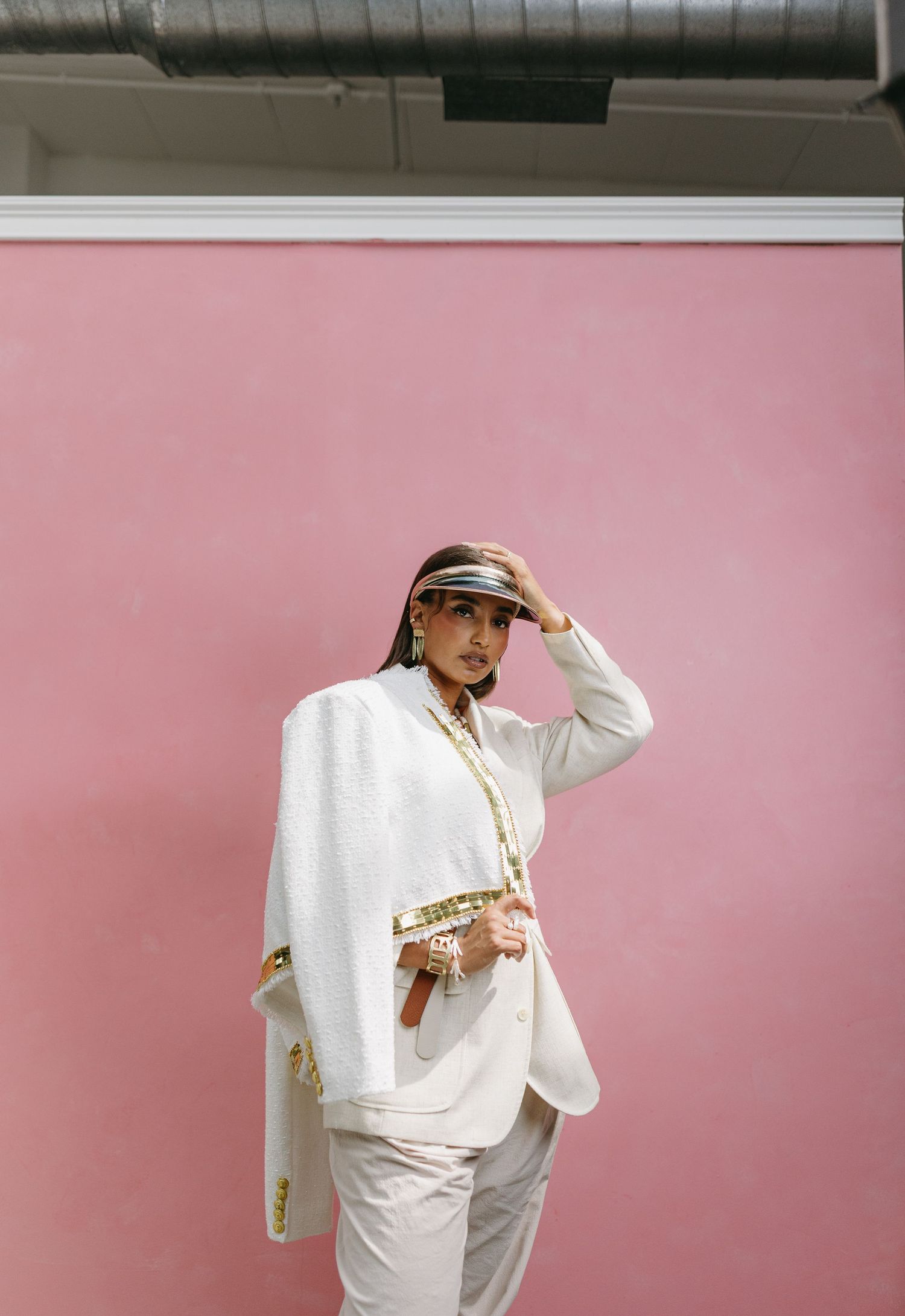 Person in white suit and gold accessories poses confidently against pink wall.