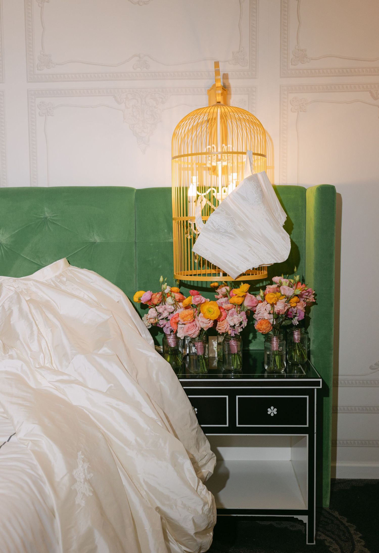 Green velvet headboard with gold birdcage lamp and colorful floral arrangement on black mirrored nightstand.