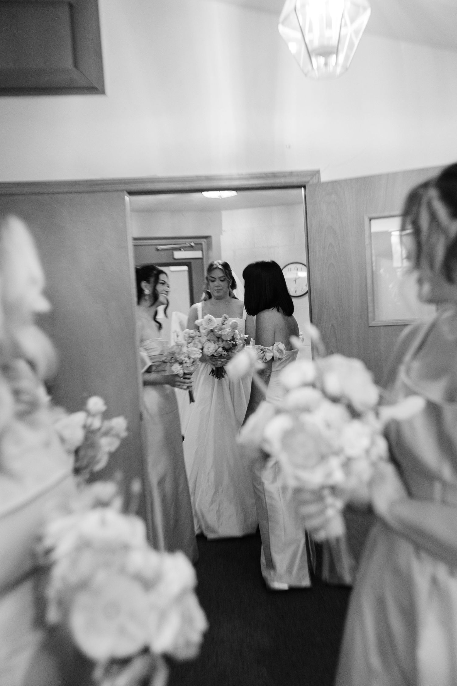 Black and white photo of bridesmaids holding bouquets reflected in a mirror while helping bride prepare.