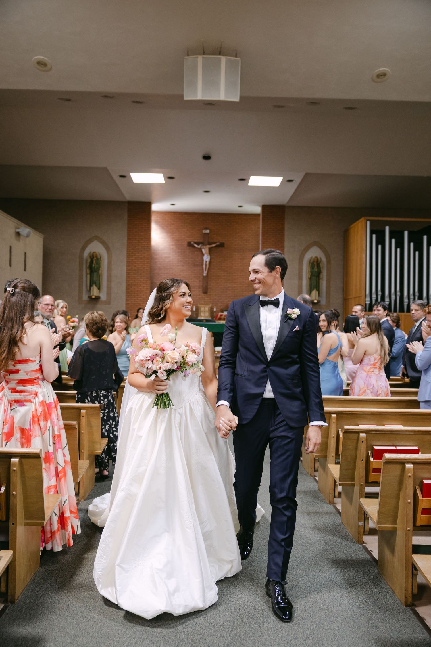 Bride and groom walk down church aisle after wedding ceremony, bride holding pink bouquet, guests applauding in pews.
