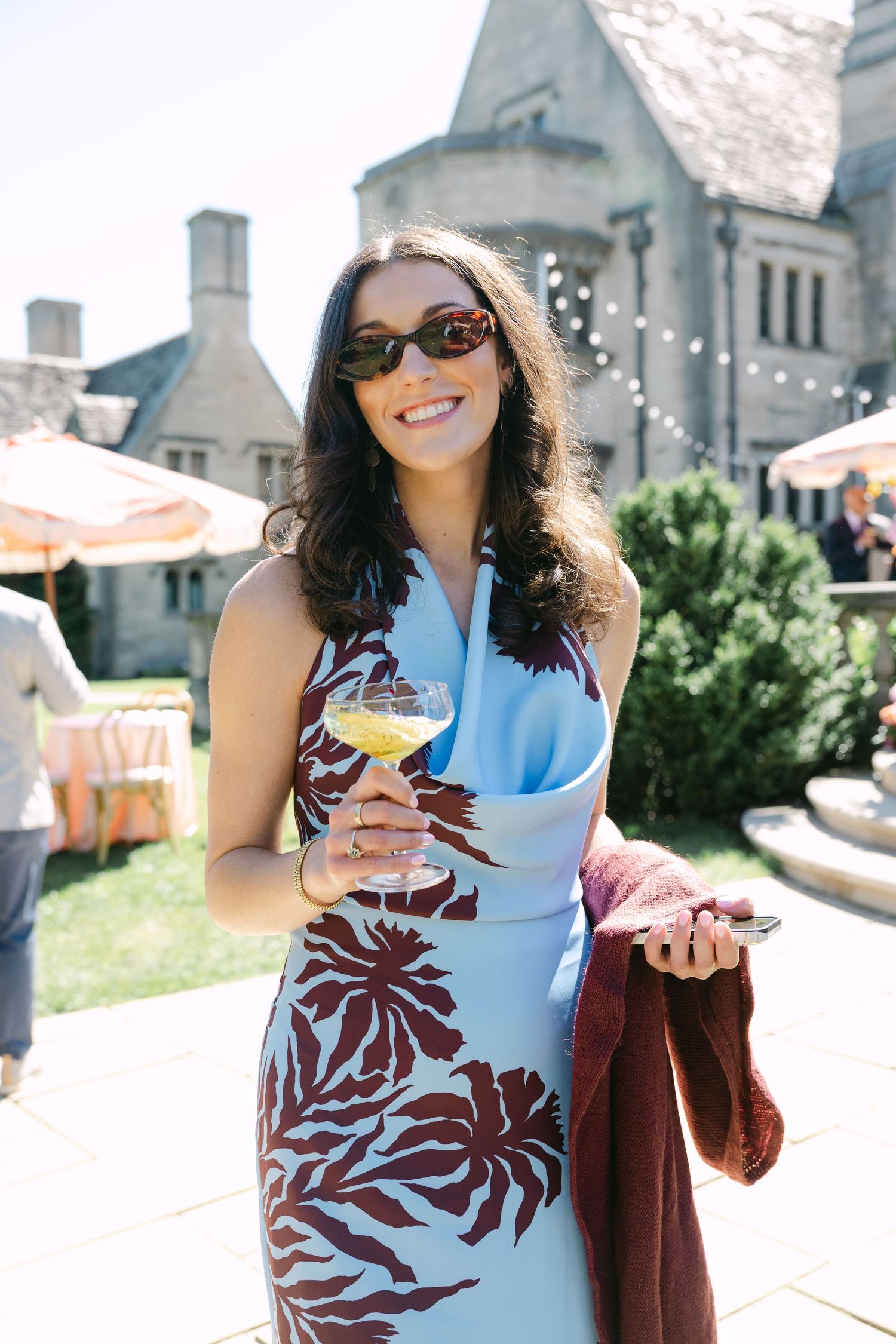A smiling woman in a blue floral dress and sunglasses holds a cocktail glass outdoors at an elegant estate event.