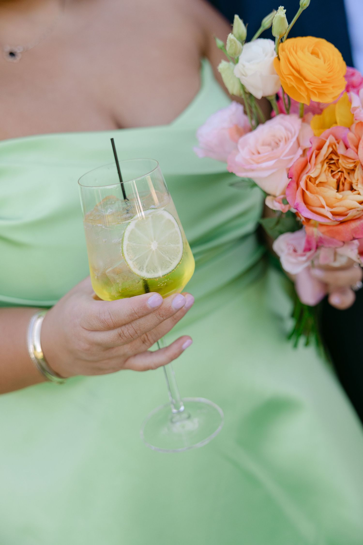Bridesmaid in mint green dress holding cocktail with lime slice near colorful floral bouquet.