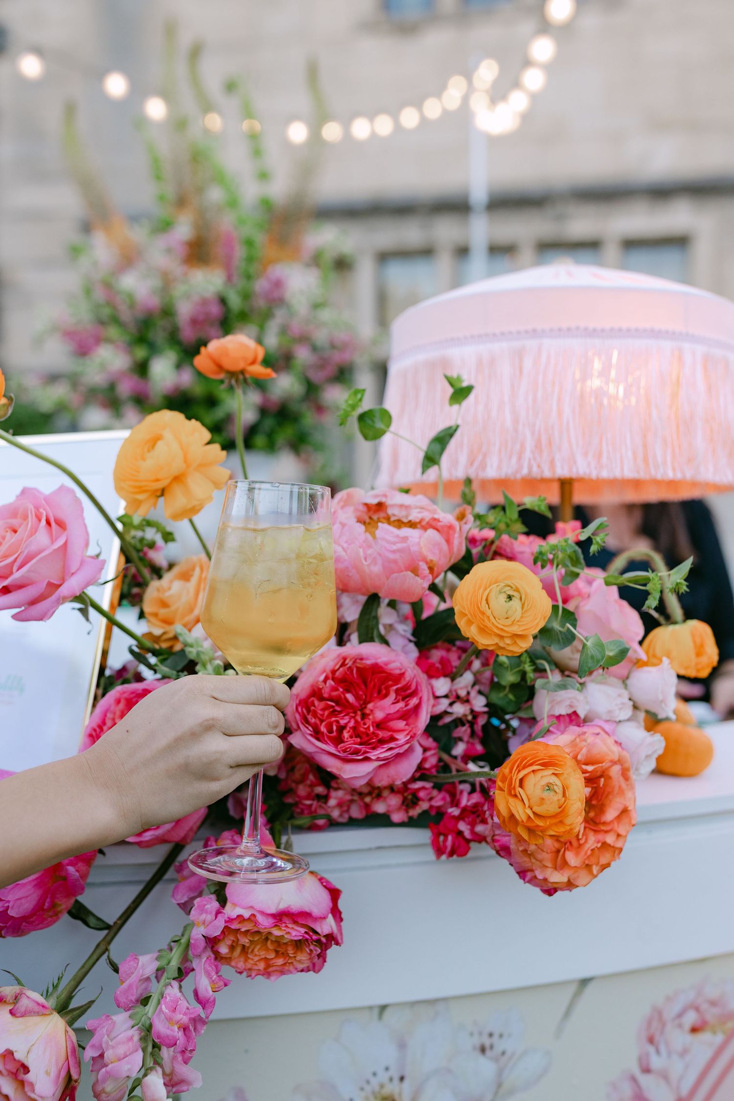 Hand holding champagne flute surrounded by vibrant pink and orange roses at outdoor wedding bar.