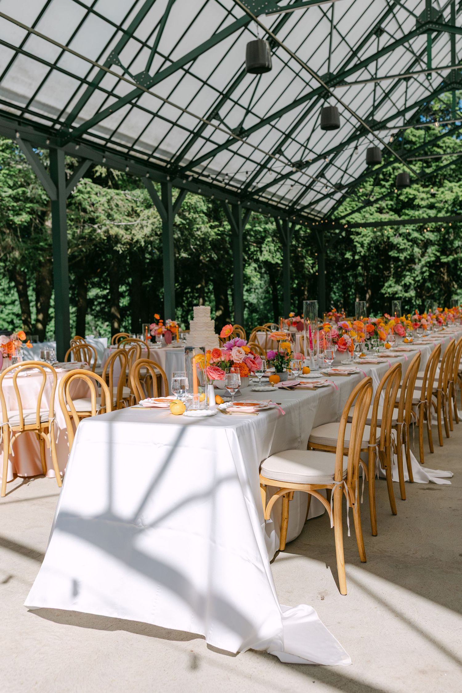 Elegant wedding reception under glass greenhouse roof with long tables, white linens, and vibrant orange and pink floral centerpieces.