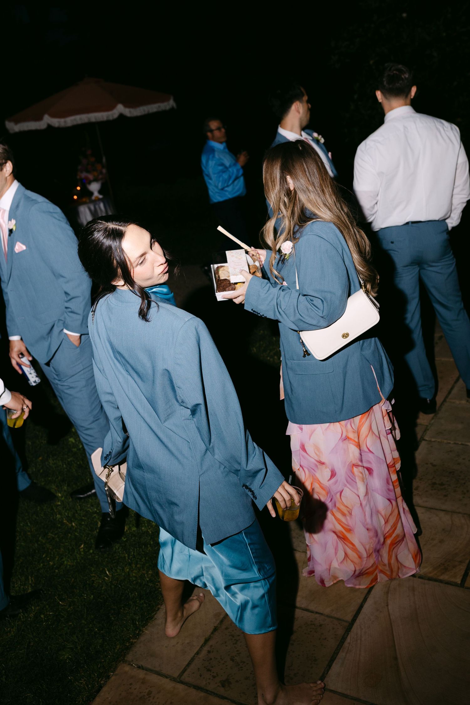 Wedding guests in blue jackets socializing outdoors at night, one holding a dessert cup and chopsticks.