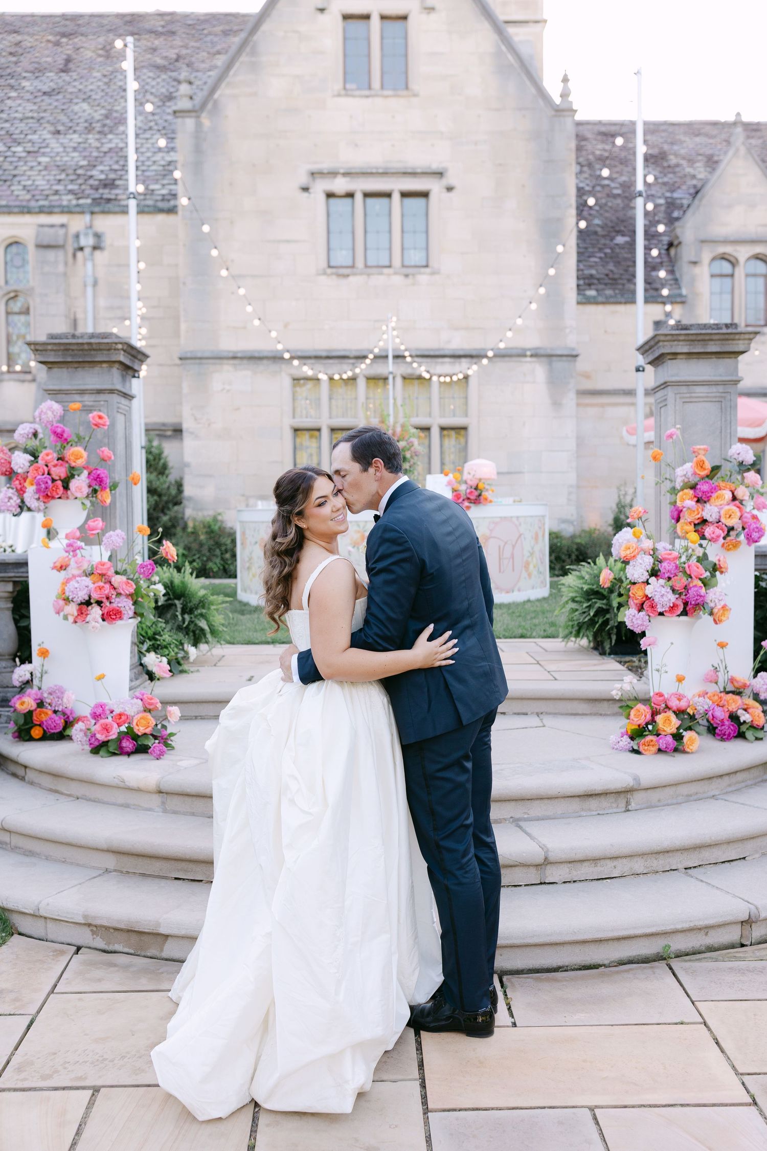 Bride and groom embrace at sunset in front of stone manor with colorful floral arrangements and wedding cakes.