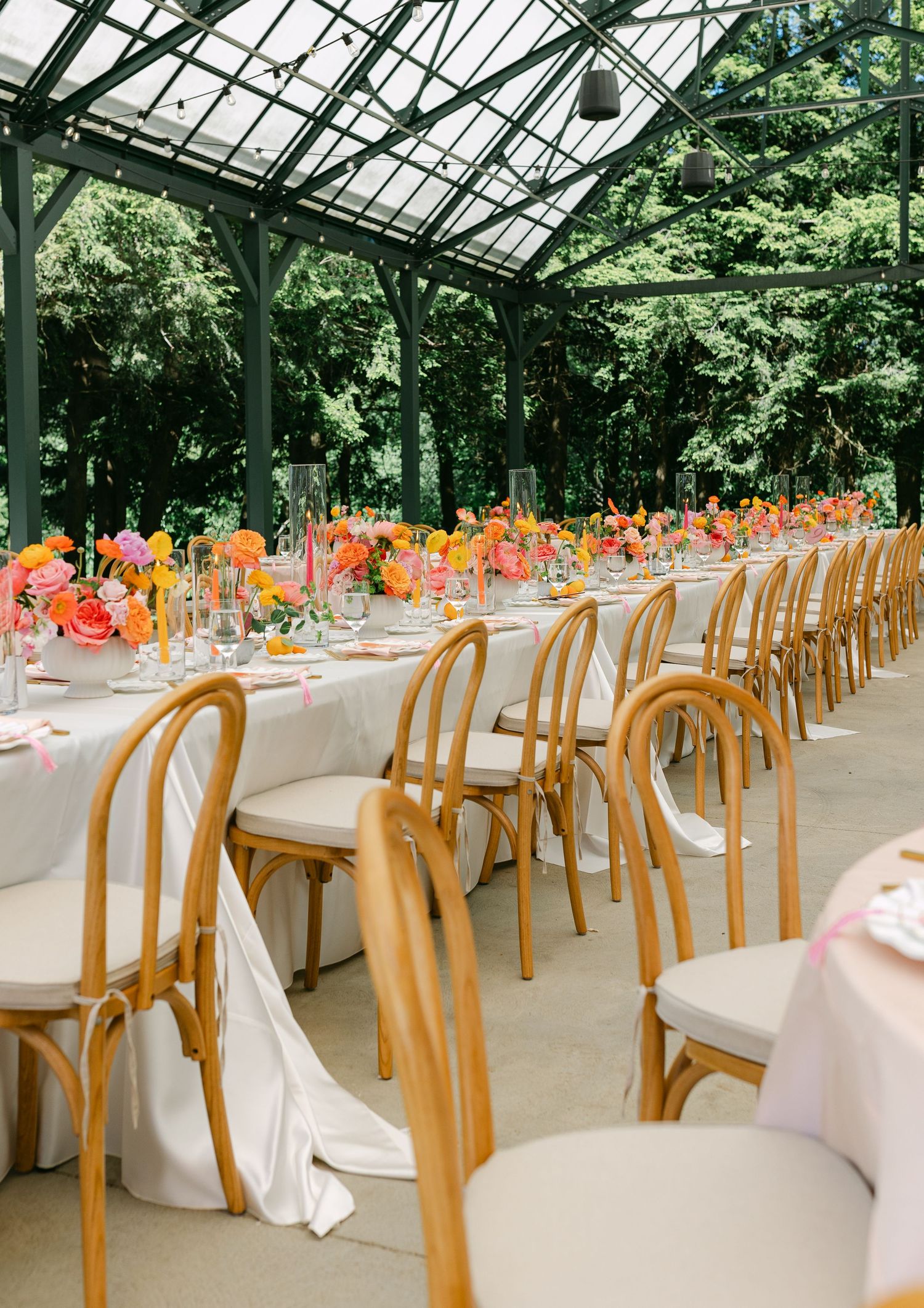 Greenhouse wedding reception with long tables, wooden chairs, and vibrant orange and pink floral centerpieces.