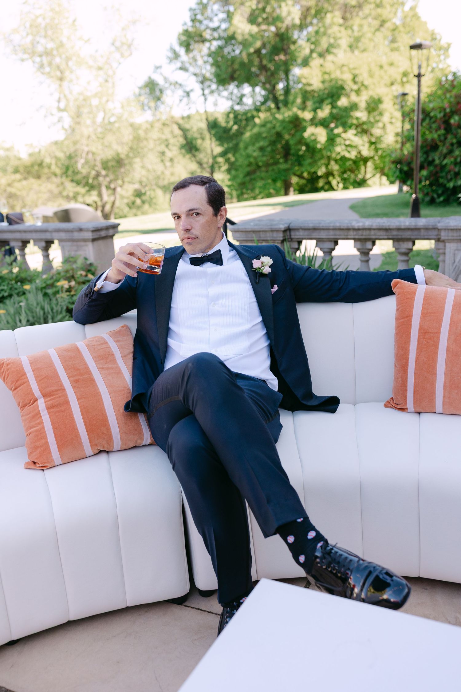 Stylish groom in navy tuxedo with bow tie relaxing on white outdoor sofa, holding whiskey glass at wedding reception.
